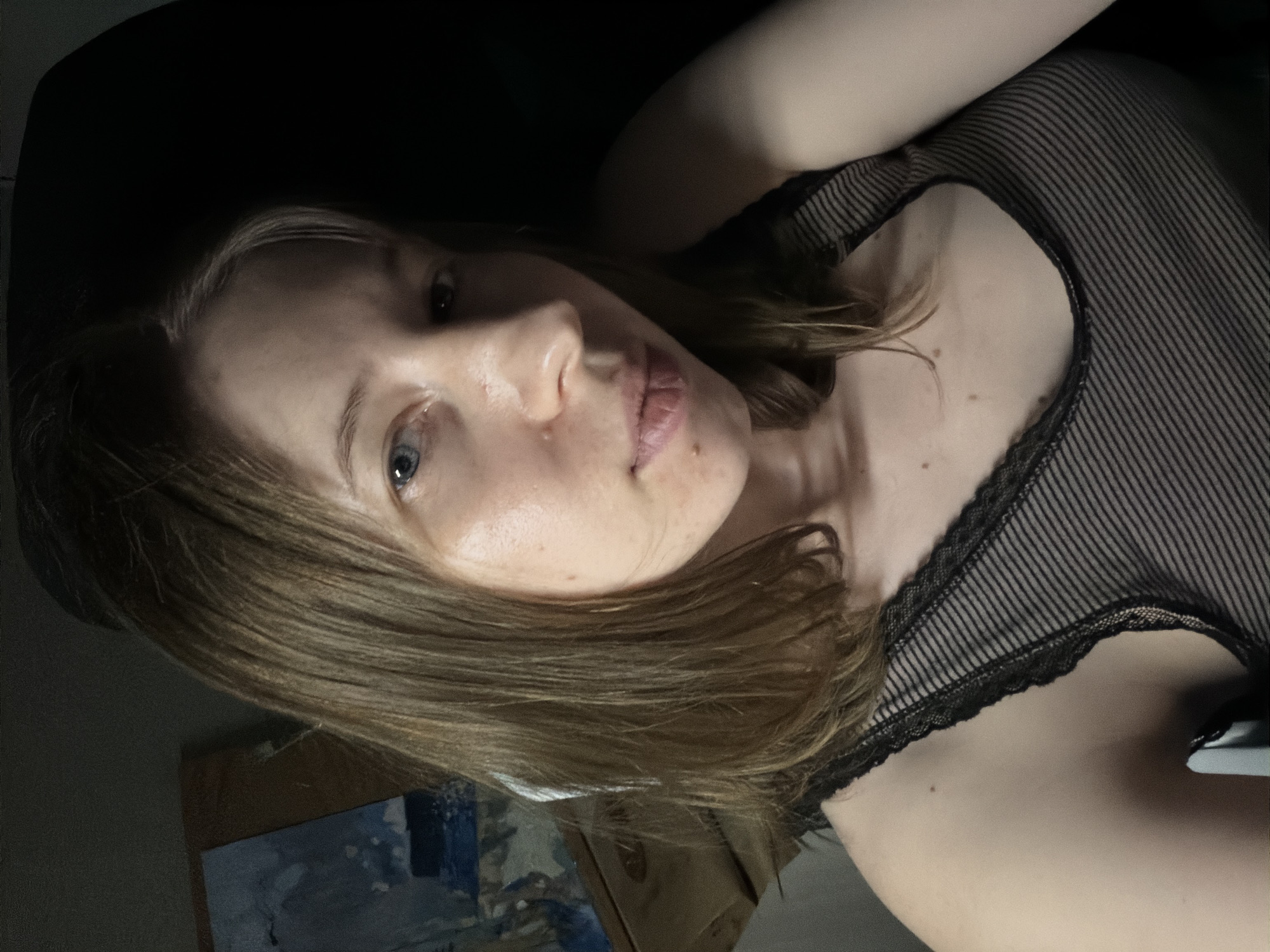 A young woman with shoulder-length, light brown hair is pictured in a close-up, self-taken photograph. She is looking directly at the camera with blue eyes and has a fair complexion. Her lips are slightly parted, and there is a small mole on her chin. She is wearing a black, sheer, striped top with lace detailing on the straps. The background is dark and out of focus, with a hint of a painting on a wooden surface to her left. The lighting is dim, casting shadows on her face and neck, suggesting an indoor setting at night. The image does not provide any specific information about the location in Dongducheon-si, South Korea.