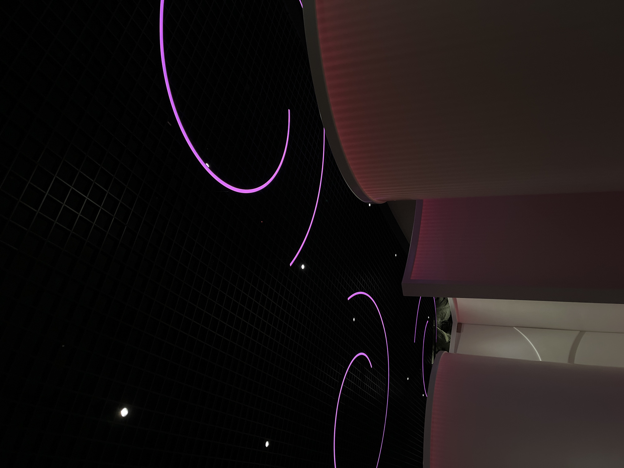This image captures a striking, abstract view of a modern architectural interior, likely an art installation or a contemporary public space, located in Krasnodar, Russia. The composition is dynamic, presenting a tilted perspective of contrasting light and shadow.
The dominant feature on the left and center is a large, dark surface, possibly a wall or ceiling, subtly textured with a grid-like pattern. Against this dark backdrop, several glowing purple arcs and curves stand out vibrantly. These are likely neon lights or LED strips, creating a sense of movement and depth. Interspersed among these purple lights are small, scattered white dots, which could be miniature light sources or reflections.
On the right side of the frame, large, curvilinear architectural elements in shades of off-white and light beige are visible. These structures appear layered and substantial, potentially forming part of a ceiling or an upper wall section. They exhibit a finely ribbed or corrugated texture. A soft pinkish-purple glow is cast upon some of these structures, likely a reflection from the bright purple lights on the opposite side, enhancing the cohesive color scheme of the space.
There are no people or discernible activities taking place, placing the focus entirely on the interplay of light, form, and texture. The scene suggests an artificially lit environment, characteristic of an interior at night or a space designed to control its illumination. The overall mood is modern, artistic, and somewhat futuristic. No text is visible within the image.