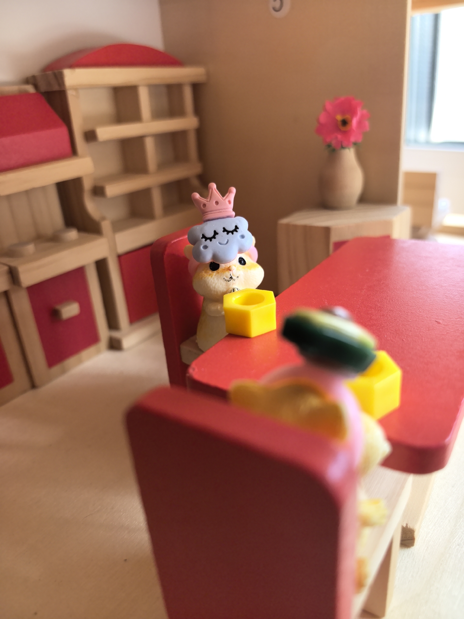 The image depicts a miniature dollhouse interior, possibly a dining room scene. On a red table, two toy figures are seated at red chairs. One figure features a light-blue cloud with a pink crown, stacked atop a yellow character. A yellow cube rests in front of the figure. The other figure, partially blurred, has green and yellow components. The table and chairs are wooden. In the background, there's a wooden kitchen cabinet with red doors and a flower in a vase. A window provides natural lighting. The environment seems to be a staged scene rather than a real-life location. There is no indication of the city Фролы, Russia.