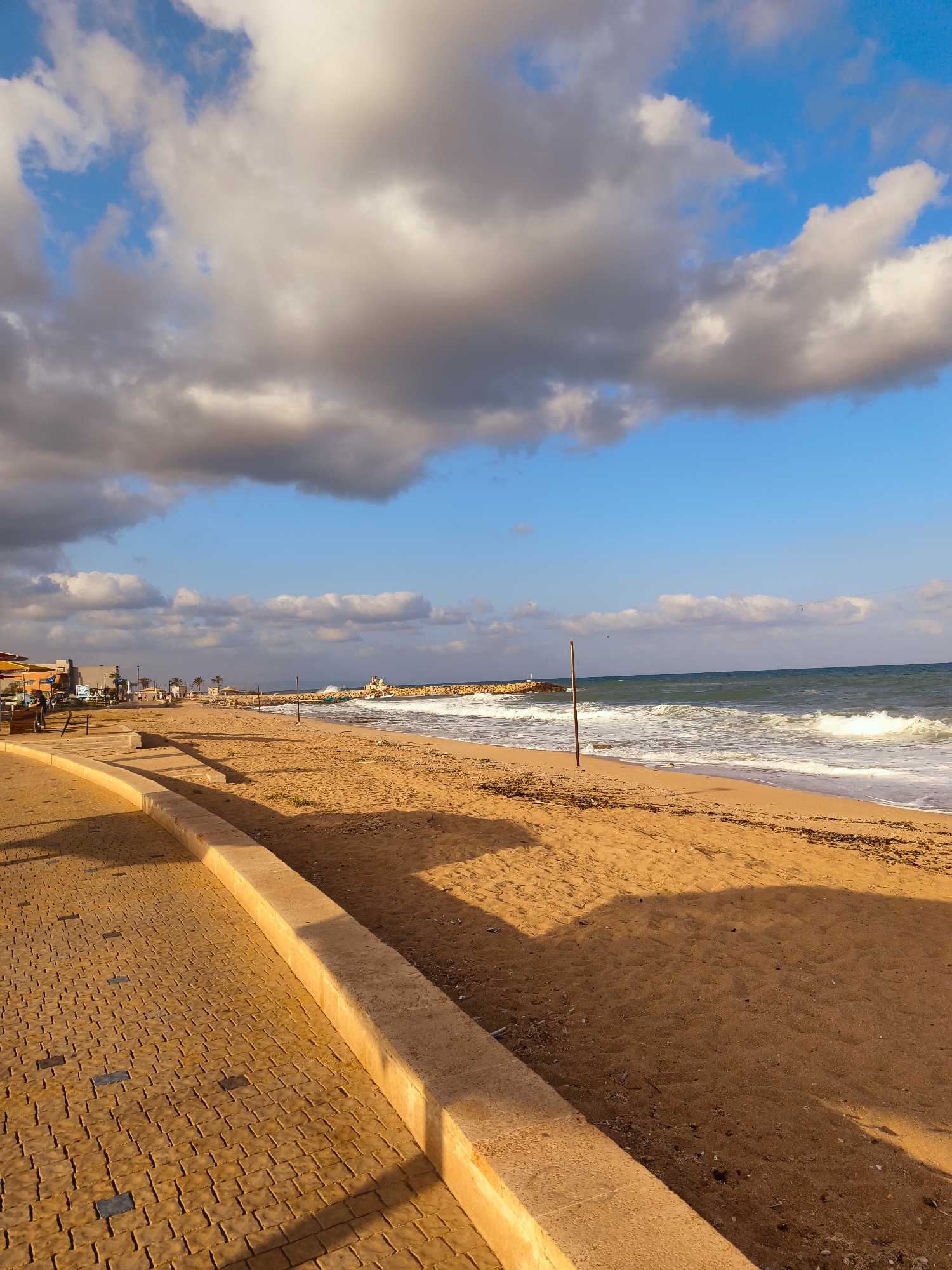 The image depicts a sunny day at a beach in Nahariyya, Israel. The foreground features a paved walkway with a textured, brick-like pattern in shades of orange and yellow, bordered by a curved concrete barrier. Beyond the barrier lies a sandy beach, with a few scattered pieces of dried seaweed. The Mediterranean Sea stretches to the horizon, with moderate waves breaking onto the shore, creating white foam. A tall, thin wooden pole stands on the sand near the water's edge. In the distance, a stone pier extends into the sea, with a lighthouse or a small structure visible at its end. Along the shoreline, on the left side of the image, there are some buildings, possibly shops or restaurants, with awnings. A few palm trees are also visible in this area, lining the promenade. The sky is a vibrant blue, punctuated by scattered white and grey clouds. The lighting suggests it is either morning or late afternoon, with long shadows cast across the sand and walkway. No people are clearly visible in the image.