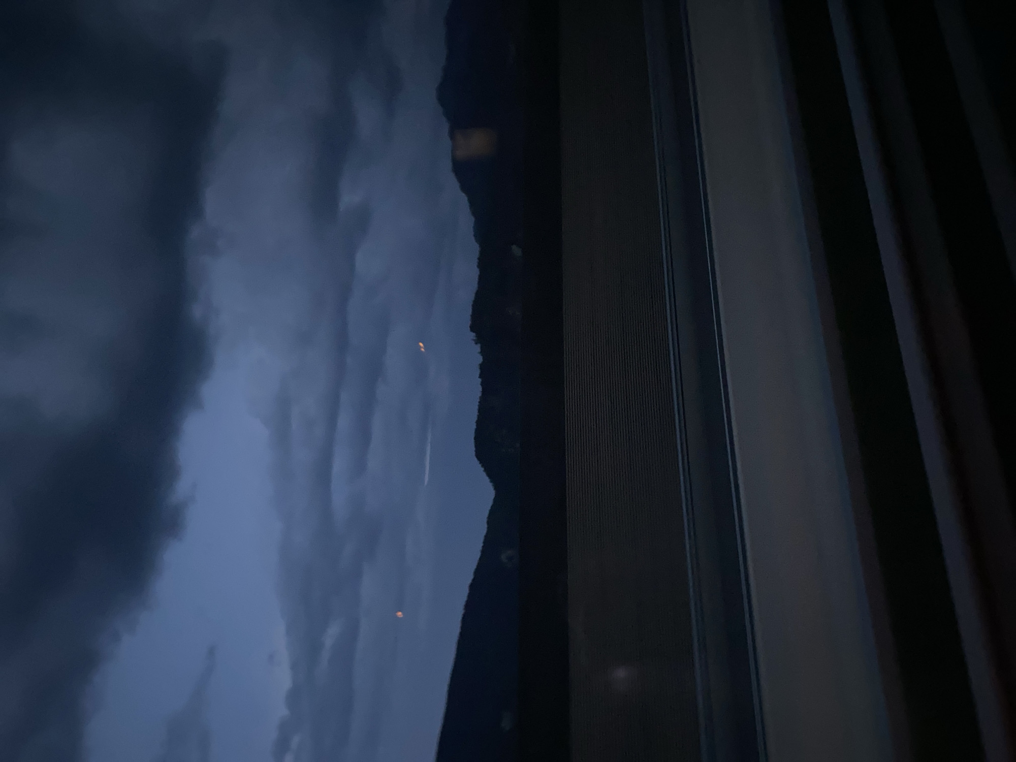 The image depicts a twilight scene with a dark, cloudy sky over a mountainous landscape. In the foreground, there appears to be a road or pathway with layered dark surfaces. The sky is filled with heavy, dark clouds, obscuring any clear view of celestial bodies except for two faint, reddish-orange circular shapes that could be distant lights or possibly the moon partially visible through the clouds. The mountains in the background are silhouetted against the dim sky. The overall impression is one of dusk or early evening, with a somewhat somber atmosphere. The location is identified as Kundl, Austria.