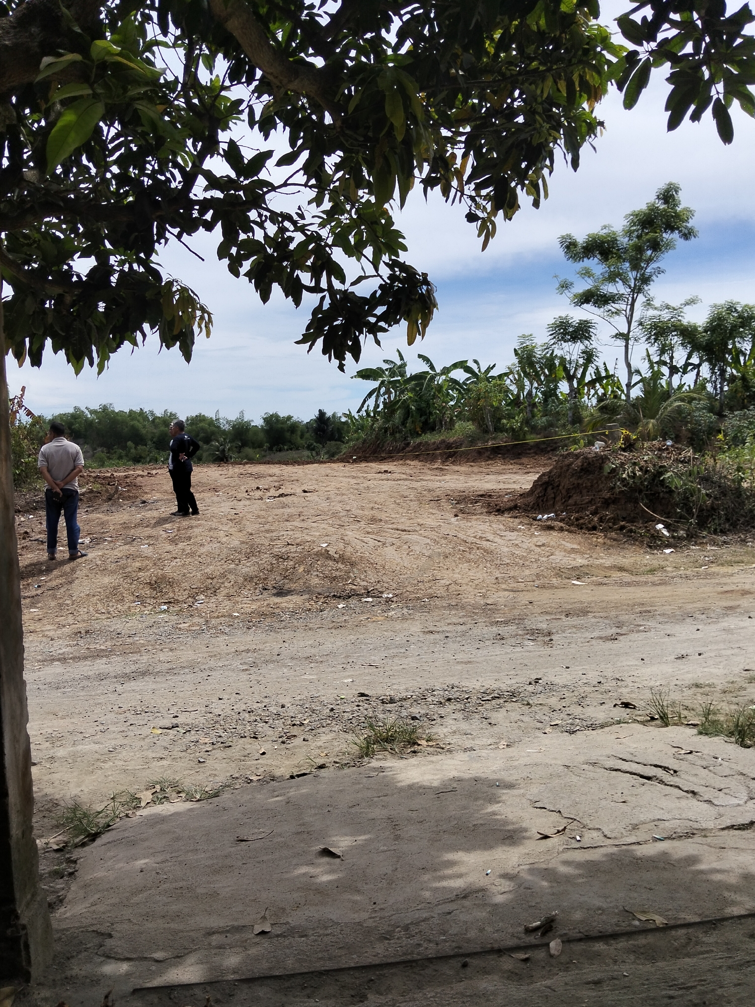 A ground-level, outdoor, daytime image captures a vacant lot in Payagaboh, Indonesia. Two individuals stand in the sandy expanse. One, wearing a light-colored polo shirt and jeans, faces away from the camera, his hands clasped behind his back. To his right, a second individual, dressed in a dark shirt and black pants, also faces away, with hands on his hips.
The terrain is mostly bare dirt and sand, with scattered debris. A pile of dark soil and vegetation sits in the middle ground, marked by a yellow caution tape stretched across the area. A line of trees and dense foliage forms the background, suggesting a rural or semi-rural setting. Overhanging tree branches with green leaves frame the top of the image. The sky above is partly cloudy with a bright blue expanse. The foreground consists of a cracked concrete surface with sparse grass.