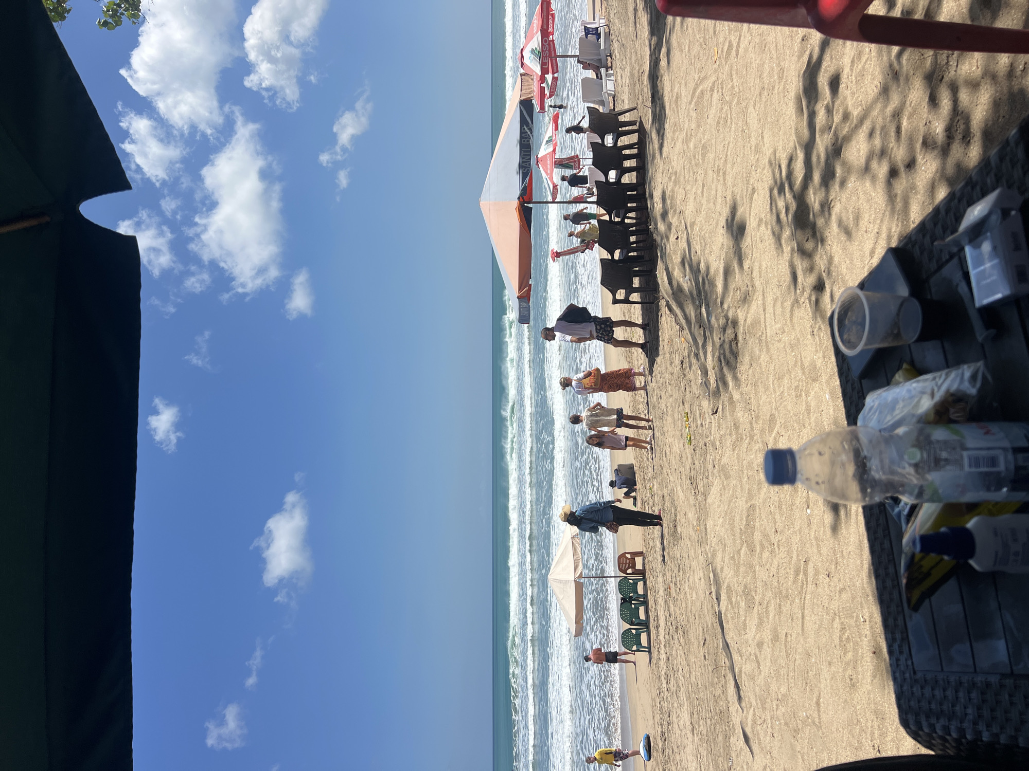 A sunny day at Kuta Beach, Indonesia, shows people enjoying the sand and sea. In the foreground, a black woven table holds a clear plastic bottle of water, a small bottle of what appears to be sunscreen, a clear plastic cup with a dark liquid, a smartphone, and a cigarette pack. Further back, several beach umbrellas provide shade. One large umbrella has an orange and white pattern, while two smaller red and white umbrellas are branded with "Santi Bar" and "Lesso." A row of black plastic chairs sits beneath the large umbrella, and white lounge chairs are positioned near the water.
Several groups of people are visible on the sand. On the left, a person in a yellow shirt stands near a blue inflatable. Near the water, a small group including children, two women, and a man are gathered. A woman wearing a straw hat stands near a single orange chair and four green chairs under a beige umbrella. Further down the beach, a man in a white shirt and shorts stands with a backpack, and a woman in a patterned skirt is beside him. More people are seen closer to the water, some wading or swimming. The ocean has moderate waves rolling onto the shore, under a bright blue sky with scattered white clouds. The sand is light brown, with some shadows cast by umbrellas and foliage from unseen trees.