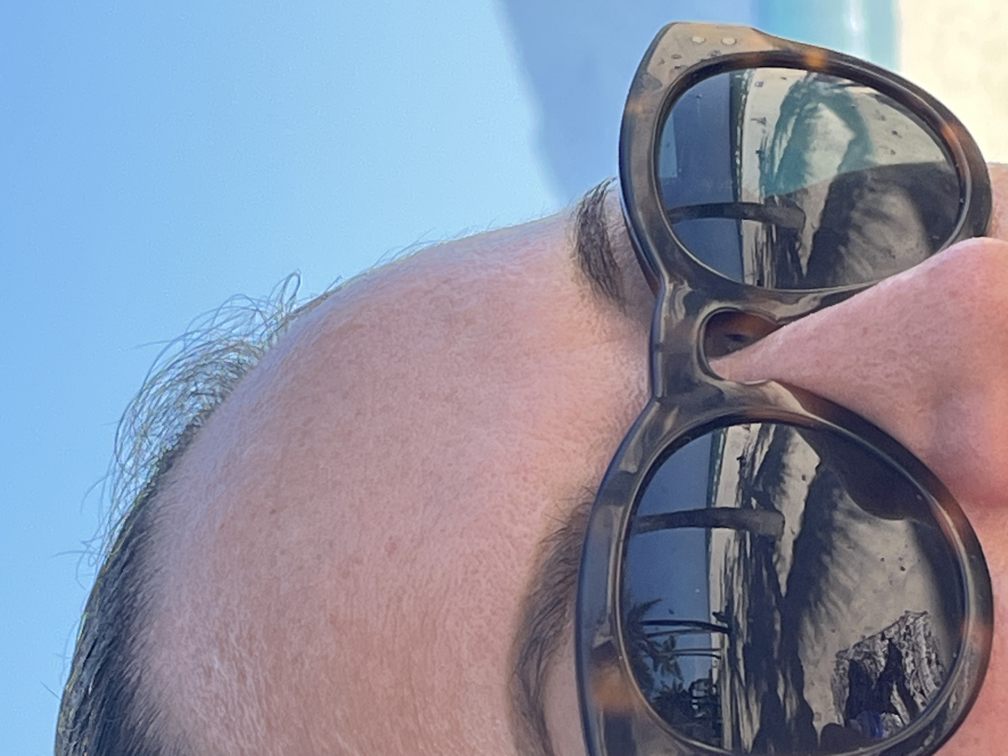 The image is a close-up shot of a person's face, focusing primarily on the forehead, hairline, nose, and a pair of tortoiseshell sunglasses. The sunglasses reflect a scene of palm trees and a building, suggesting a tropical location. Due to the reflections, it is difficult to pinpoint the exact location, but the presence of palm trees and what appears to be a building suggests a coastal or urban area. Đà Nẵng, Vietnam, is a coastal city known for its beaches and modern architecture, which fits with the imagery in the reflection.