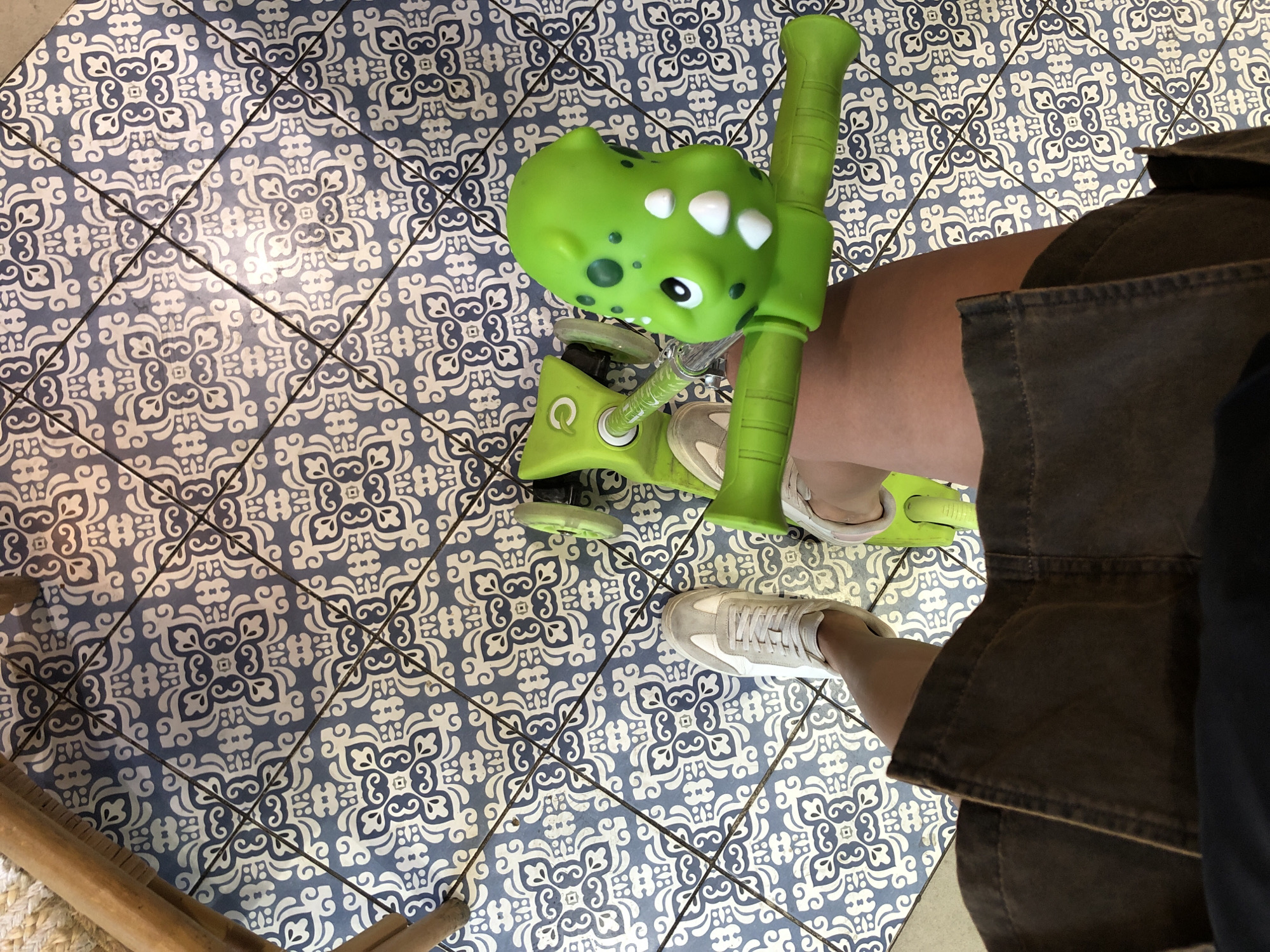 A high-angle shot shows a person's legs and feet, along with a green toy scooter, on a tiled floor. The tiles have a blue and white ornate pattern. The scooter has a dinosaur-shaped front with large, white, oval-shaped eyes. The person is wearing white sneakers and dark-colored pants. One foot is on the scooter, while the other is resting on the ground. The lighting suggests it could be daytime, possibly indoors given the tiled floor which is common in Spanish locations like València. The overall scene appears casual and relaxed, possibly a child playing or an adult with a toy. There is no visible text in the image.