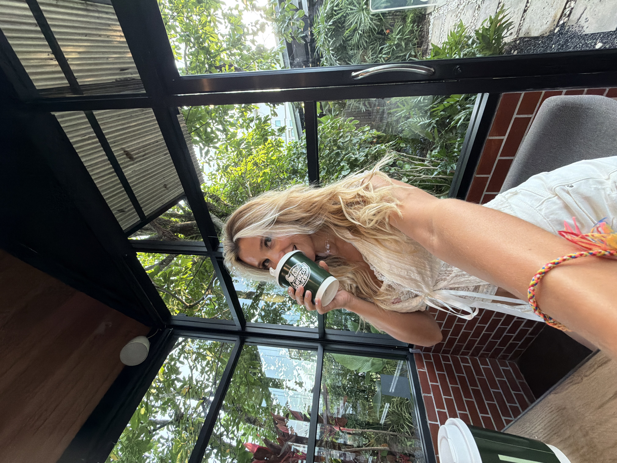 A woman with blonde hair is seated inside a glass-enclosed establishment, holding a dark green paper cup to her lips. She is wearing a white, lacy, tie-front top and white shorts. The establishment appears to be a cafe, with exposed brick walls and a wooden and corrugated metal ceiling. Outside the windows, lush green foliage and trees are visible, suggesting a tropical environment, consistent with the location of Sakhu, Thailand. The lighting indicates it is daytime, possibly late afternoon, with sunlight filtering through the leaves. A colorful woven bracelet is visible on the woman's wrist. A portion of another cup and arm are also in the foreground, suggesting the photo is a selfie.