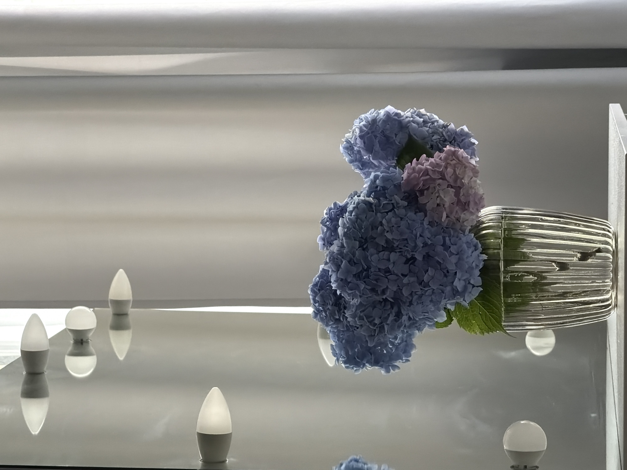 The image shows a still life arrangement on a reflective surface, likely a table or countertop. The primary subjects are a bouquet of blue and pink hydrangeas in a ribbed, metallic vase, and several small, modern LED lights scattered around the surface. The hydrangeas are the focal point, with their delicate, clustered petals dominating the right side of the frame. A few green leaves are visible amongst the flowers. The vase is cylindrical with a textured, metallic finish.
The LED lights are of two distinct types: some are elongated and tapered with a frosted white top, while one is spherical and entirely white. They are positioned at various points on the reflective surface, and their reflections are clearly visible beneath them, creating a sense of depth and symmetry. The lighting in the scene is soft and diffused, suggesting an indoor setting, possibly with ambient light from above or from the lights themselves. The overall mood is one of modern elegance and tranquility. There is no visible text in the image. The location context of Sochi, Russia, doesn't provide specific visual cues within the image itself, but suggests an urban or domestic interior setting. The time of day is indeterminate due to the artificial lighting and controlled environment.