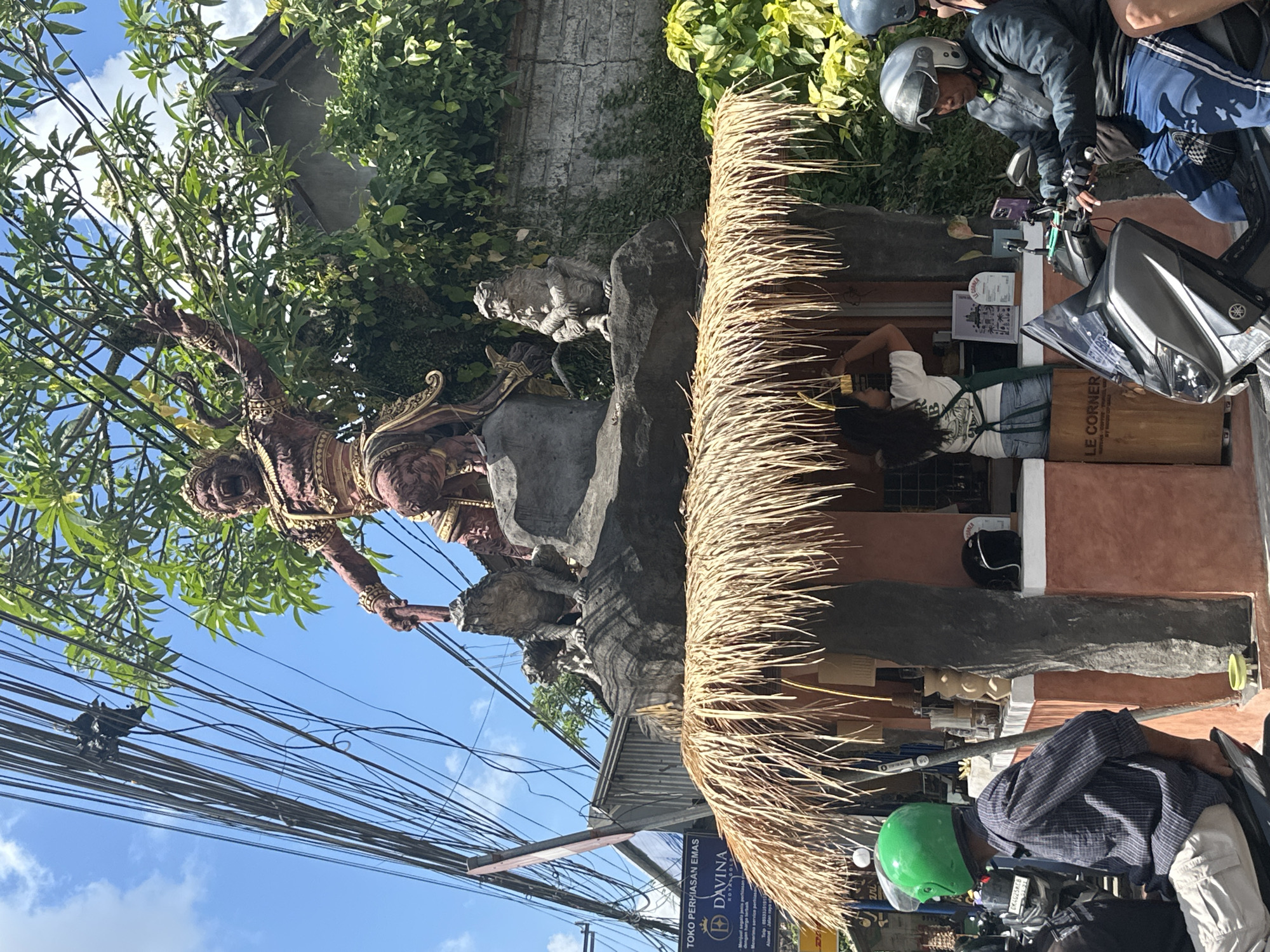 In Ubud District, Indonesia, a large statue of a mythological figure, possibly a deity or demon from Hindu mythology, stands prominently above a small open-air coffee stand. The statue, rendered in reddish-brown and gold, is in a dynamic pose, holding a staff or weapon and accompanied by two smaller statues of monkeys or similar creatures perched on rocks. The coffee stand, with a thatched straw roof, is situated at street level. A woman in an apron is visible behind the counter, serving customers. Several people on motorcycles are stopped at or passing by the stand. One person in the foreground wears a green helmet and a striped shirt. Another person on a motorcycle wears a silver helmet and a jacket. The scene is cluttered with overhead electrical wires, common in many urban and semi-urban areas. Lush green foliage surrounds the statue and the building behind the coffee stand, suggesting a tropical environment. The sky is blue with scattered clouds. A sign for "Toko Perhiasan Emas Davina Royal Gold" is visible on a blue banner in the background.