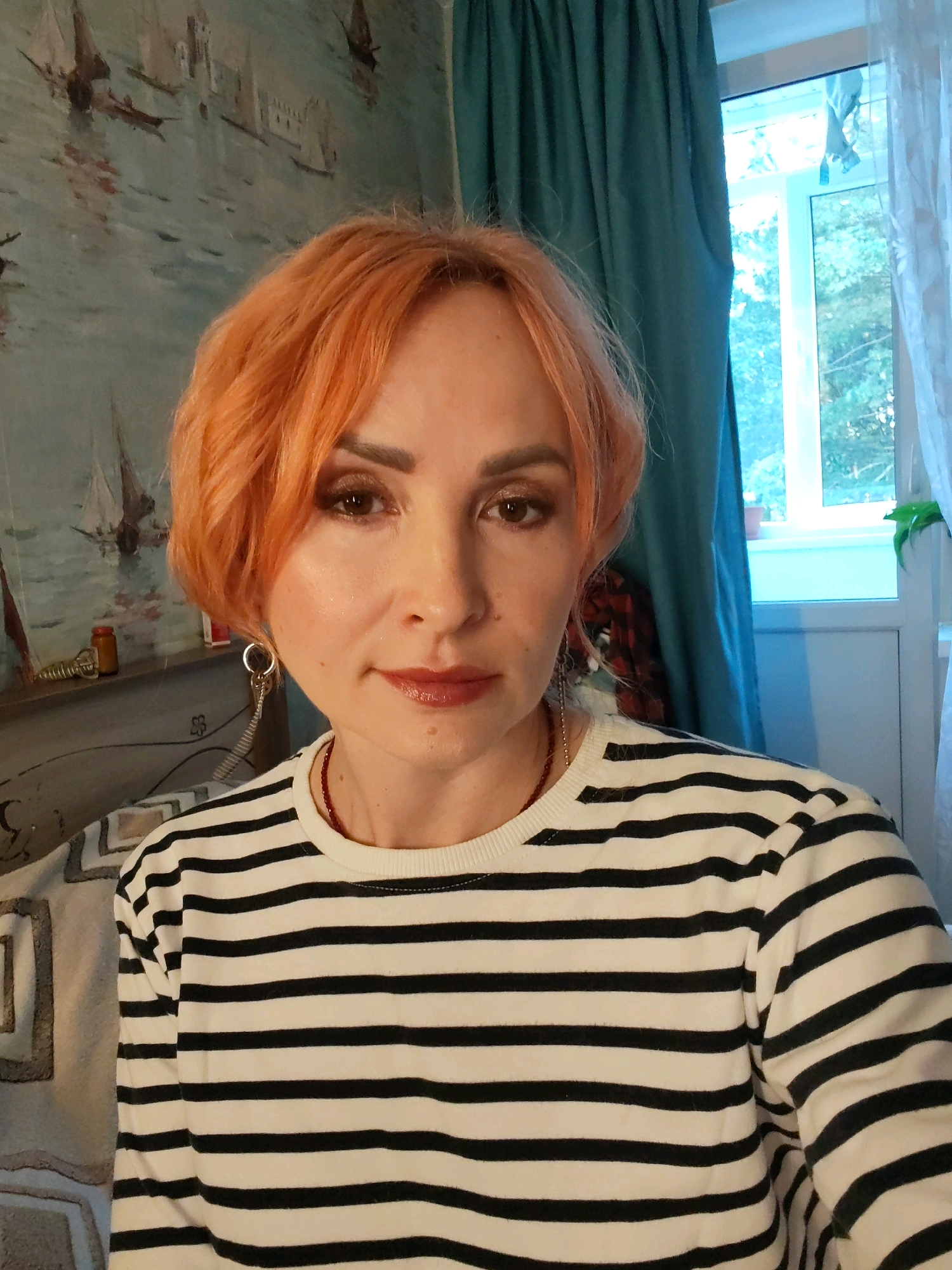 A close-up, head-and-shoulders selfie features a Caucasian woman with short, dyed orange hair and light skin. She is wearing a long-sleeved, black-and-white striped shirt and a thin red beaded necklace. Her left earlobe has a dangling silver earring with a coiled spring attached. She is wearing makeup, including eyeshadow, mascara, and lipstick. Her gaze is directed towards the camera.
The background consists of a wall with a wallpaper depicting sailing ships on a body of water. To the right of the woman, a window with white trim is visible, covered by a dark teal curtain on the left and a sheer white curtain on the right. Through the window, trees and sky are discernible. The lighting suggests an indoor setting, possibly a room in Chaikovsky, Russia, given the presence of domestic elements like curtains and wallpaper.