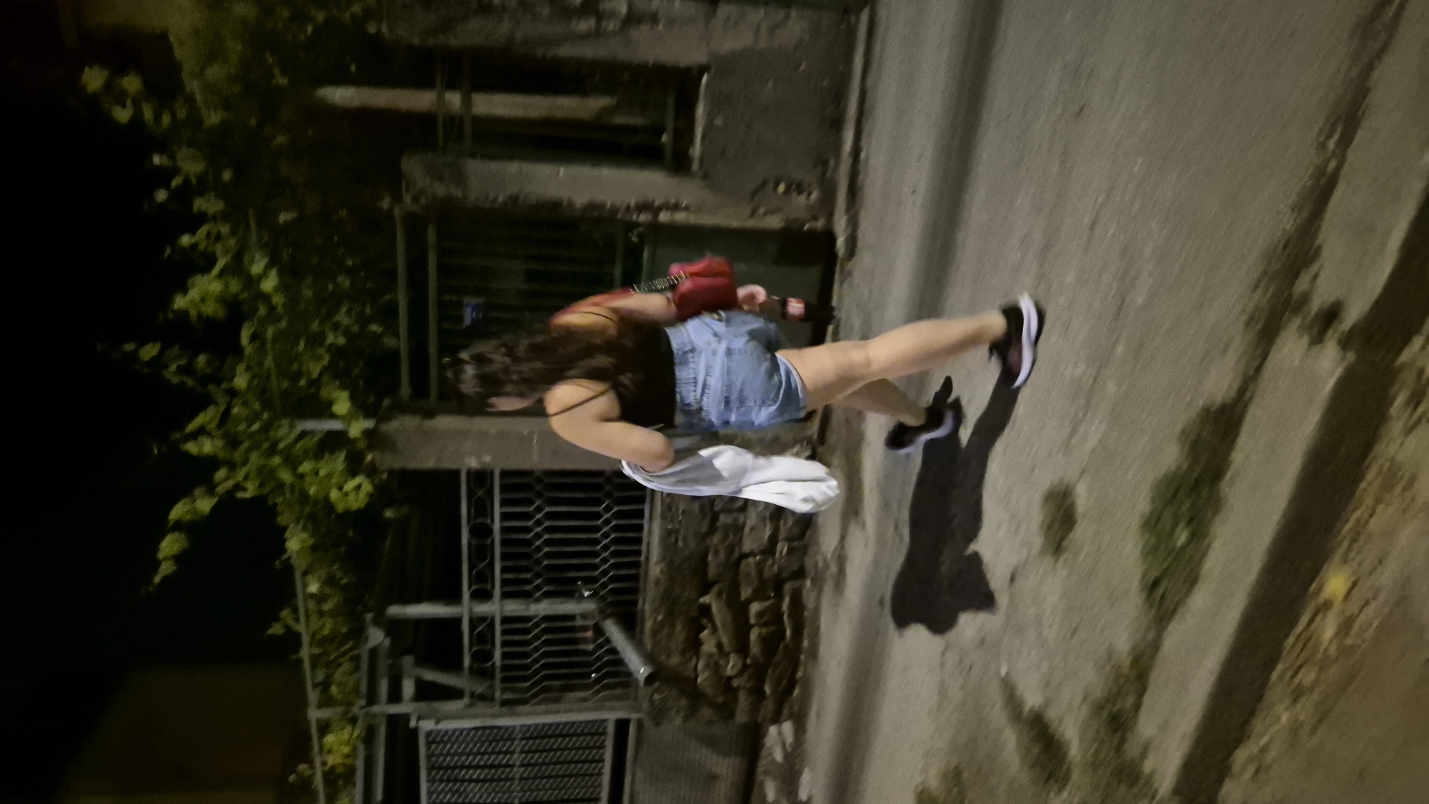 A person walks away from the camera on a street at night. The person is wearing a black tank top, denim shorts, and sneakers. They are carrying a white garment in their left hand and a red purse over their right shoulder. The street is paved and appears to be in a residential area, with a fence and buildings visible in the background. There are trees and foliage in the background, suggesting a potentially rural or suburban setting. The lighting suggests it is nighttime, with some artificial light sources present. The image does not contain specific indicators to confirm the location in Lovech, Bulgaria.