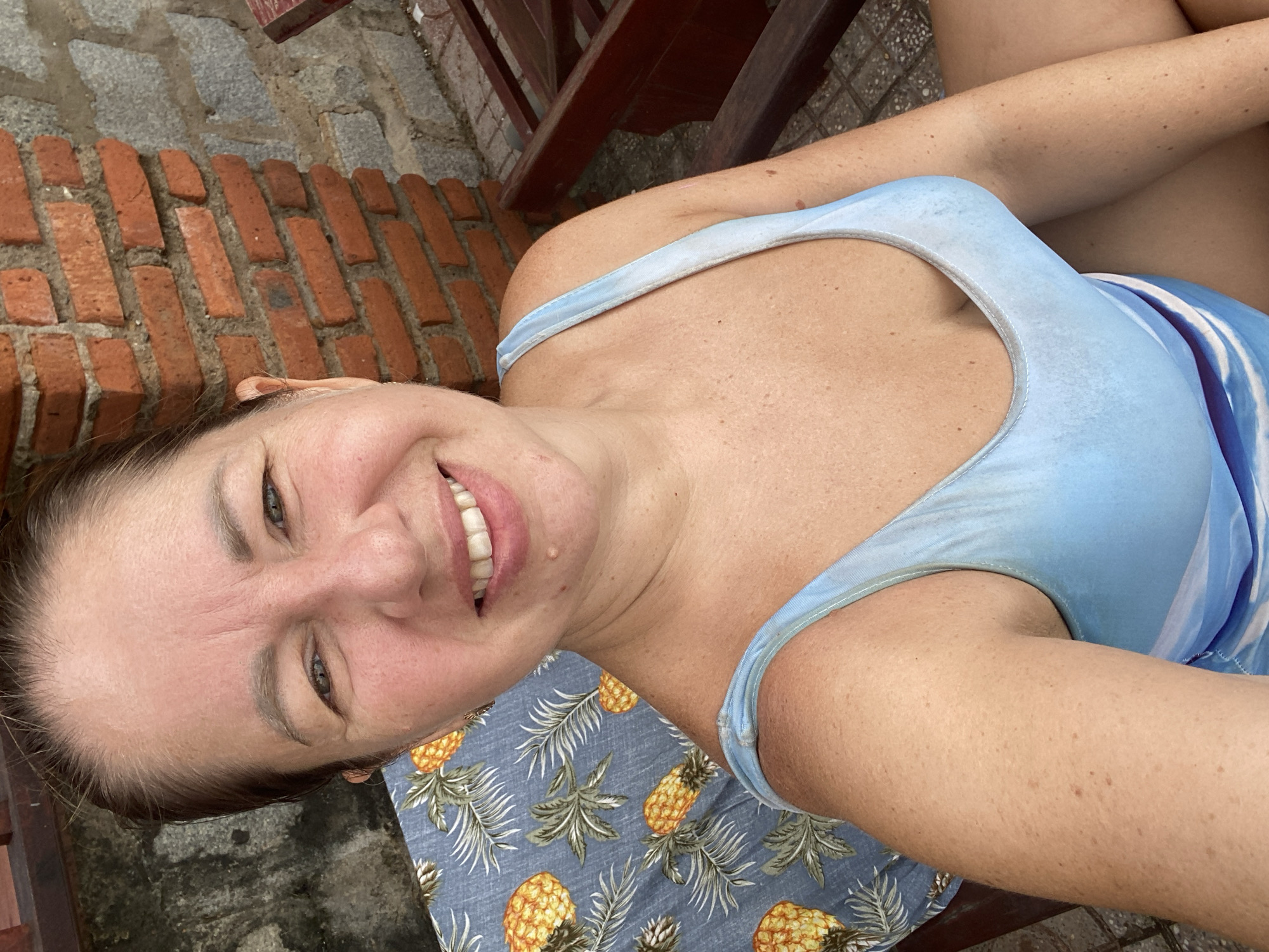 In Mui Ne, Vietnam, a medium-shot selfie captures a woman with light skin and brown hair, wearing a blue swimsuit with a pineapple print on the fabric draped behind her. She is seated on a wooden chair, with a brick wall visible in the background. The scene suggests an outdoor setting, possibly near a beach or resort.