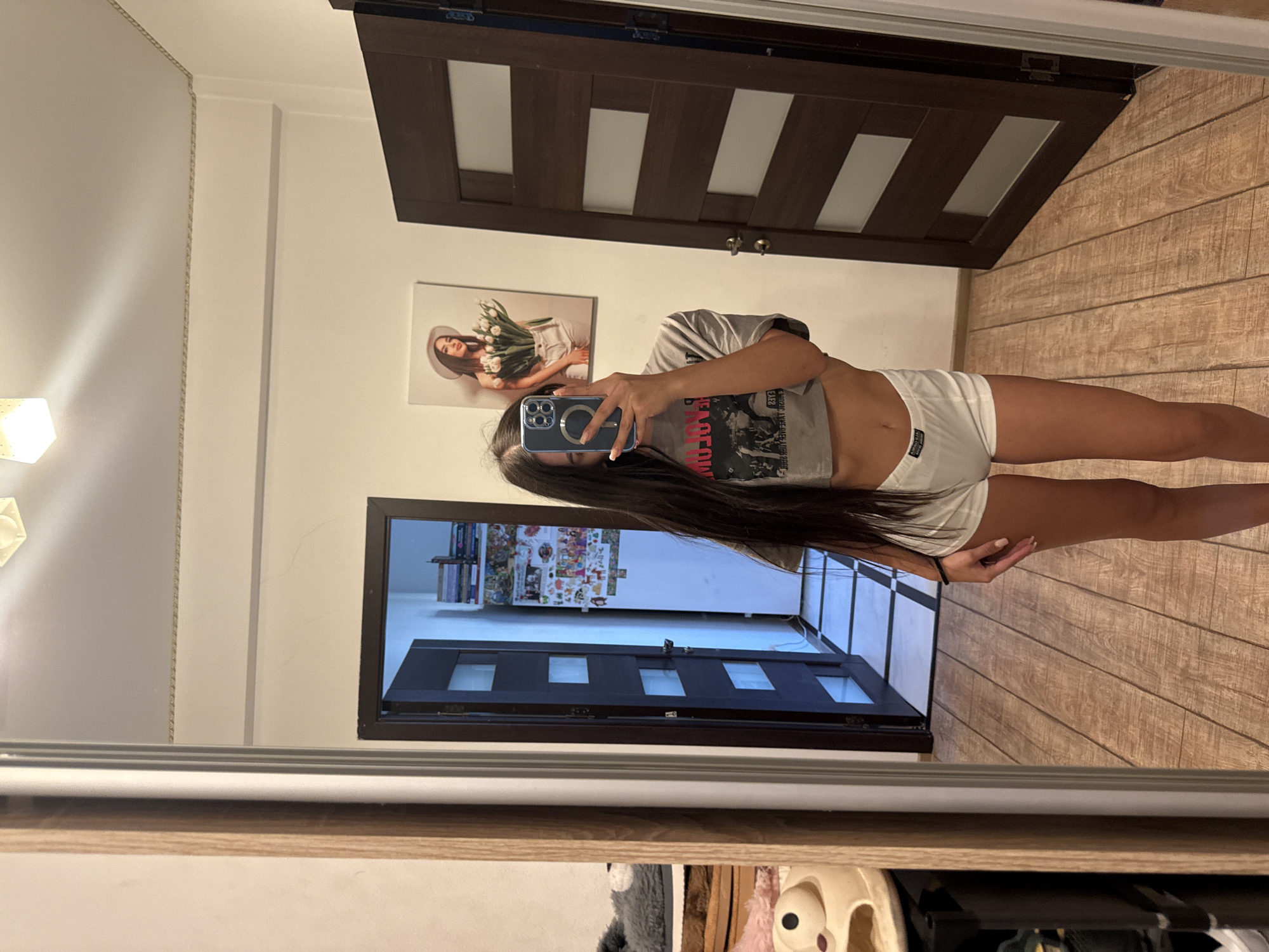 The image, taken in Chișinău, Moldova, shows a young woman with long dark hair taking a selfie in a mirror. She is wearing a grey crop top with a graphic print and white shorts. Her reflection is visible in a full-length mirror that also captures a portion of the room. Behind her, a dark brown wooden door with frosted glass panels is visible. To the left of the door, a framed picture of a woman wearing a hat and holding a bouquet of white tulips hangs on the wall. The room appears to be a bedroom or dressing area, with wooden flooring. In the foreground, the edge of a wooden shelf unit is visible, with some soft toys and other items stored on it. The lighting suggests it is daytime, but no specific time can be determined. The woman's expression is neutral as she focuses on her phone. The reflection also shows a glimpse of another door and a shelf with books and various items.