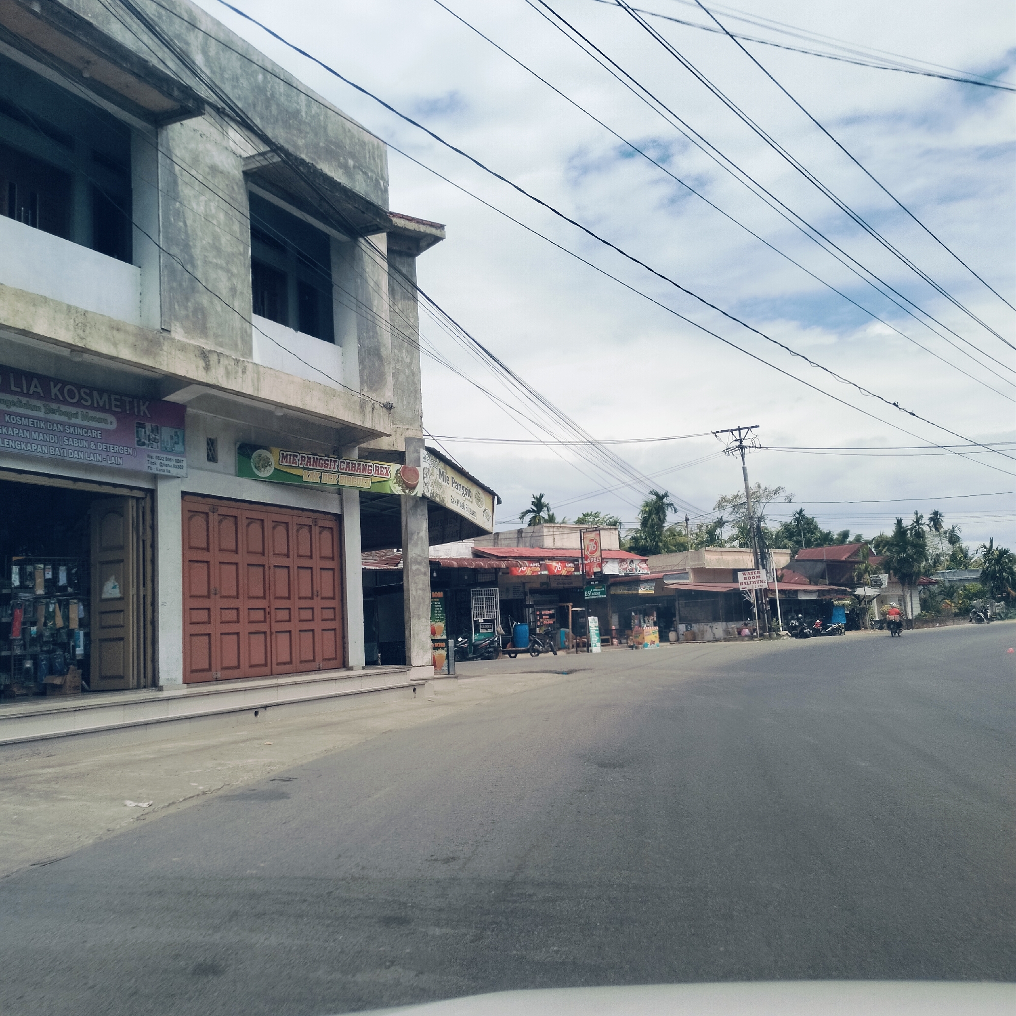 This image captures a street scene in a commercial area, likely in Keudealuerheung, Indonesia, under an overcast sky during daytime. The foreground features an asphalt road with minimal traffic.
On the left, a two-story concrete building houses businesses. The ground floor clearly displays a sign for "LIA KOSMETIK," advertising "KOSMETIK DAN SKINCARE," "PERLENGKAPAN MANDI (SABUN & DETERGEN)," and "PERLENGKAPAN BAYI DAN LAIN-LAIN." Contact details, including "Wa: 0822 8061 0887," "Tiktok: @liana.la302," and "Fb: Liana lia," are also visible. Adjacent to the cosmetics shop, a large brown folding door suggests another business entrance, above which a sign for "MIE PANGSIT CABANG REX" is displayed, advertising "Mie Pangsit."
Further down the street on the right, a series of single-story shops or stalls with various roof types line the road. Signs for "WATER ROOM HALIMUN" and a smaller "APEL" sign are discernible, along with a partially visible sign that reads "BS" (possibly for a banking agent). Several motorcycles are parked near these establishments. A few people are visible, including one person riding a motorcycle further down the road and some individuals near the stalls.
Numerous black electrical wires crisscross the frame against the cloudy sky, connected to a power pole in the mid-ground. Palm trees and other tropical foliage are visible in the background, behind the row of shops, reinforcing the Southeast Asian setting. The scene depicts typical daily commerce and local activity.