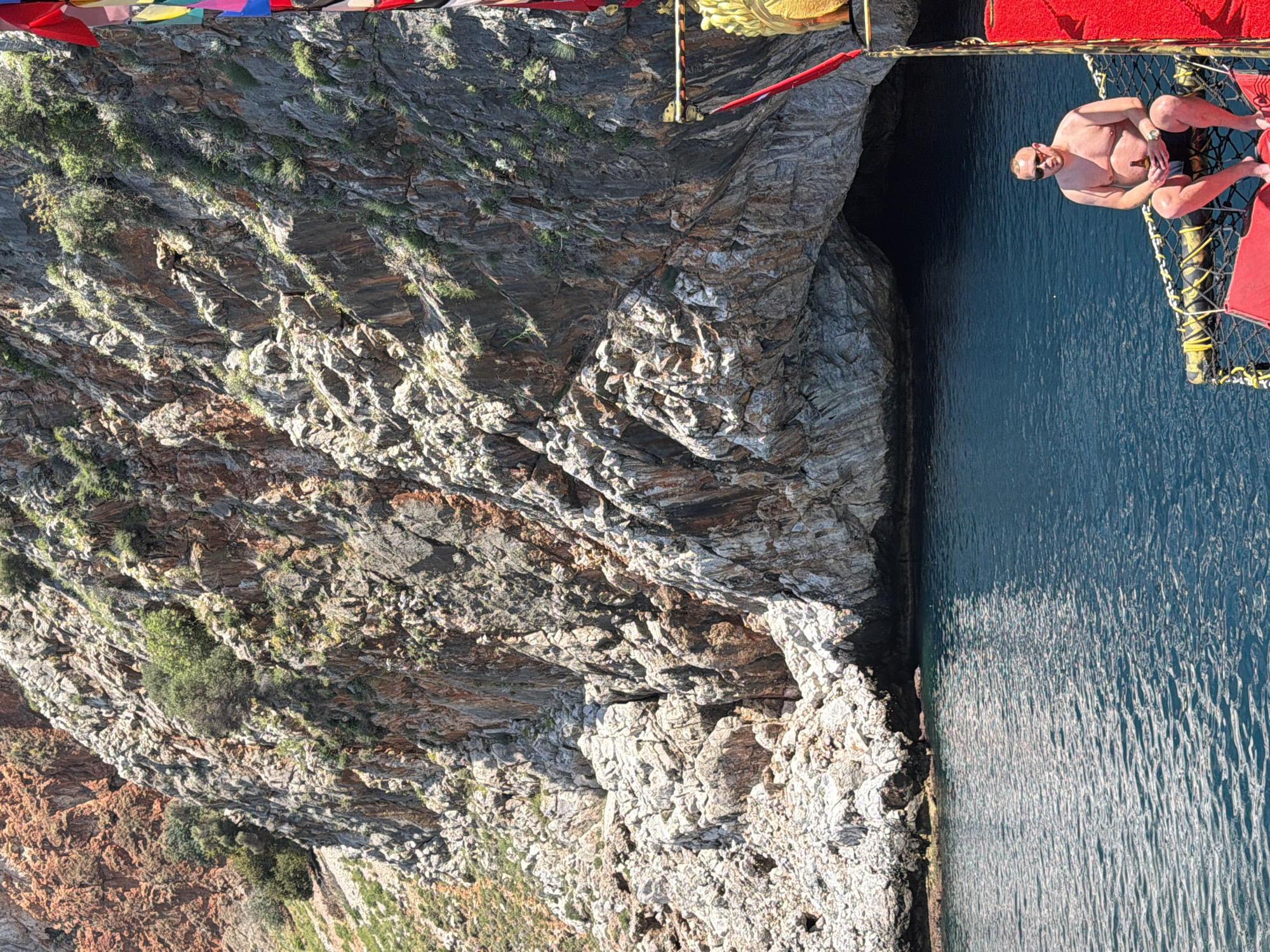 The image captures a serene coastal scene in Alanya, Turkey, on a bright, sunny day, likely during the morning or afternoon given the clear light. A man is depicted relaxing on a red-bordered net structure, possibly an extension of a boat or catamaran, positioned very close to a formidable rocky cliff face. He is shirtless, wearing sunglasses and a watch, and holds a bottle or can in his hand, appearing to be enjoying a leisure moment.
The water, a deep blue, appears calm and reflects the sunlight, creating textured patterns on its surface. The imposing cliff rises vertically from the water, displaying a rugged texture with distinct layers and variations in rock color, from dark grey to lighter brown and white. Patches of sparse green vegetation, including small bushes and grass, cling to the cliff's ledges and crevices, adding a touch of life to the otherwise stark rock.
Above the boat structure, part of a string of colorful triangular flags, resembling prayer flags, can be seen, along with what appears to be a golden, carved ornament. The overall atmosphere is one of tranquility and natural beauty, combining a leisurely activity with a stunning geological backdrop. No visible text is present in the image.