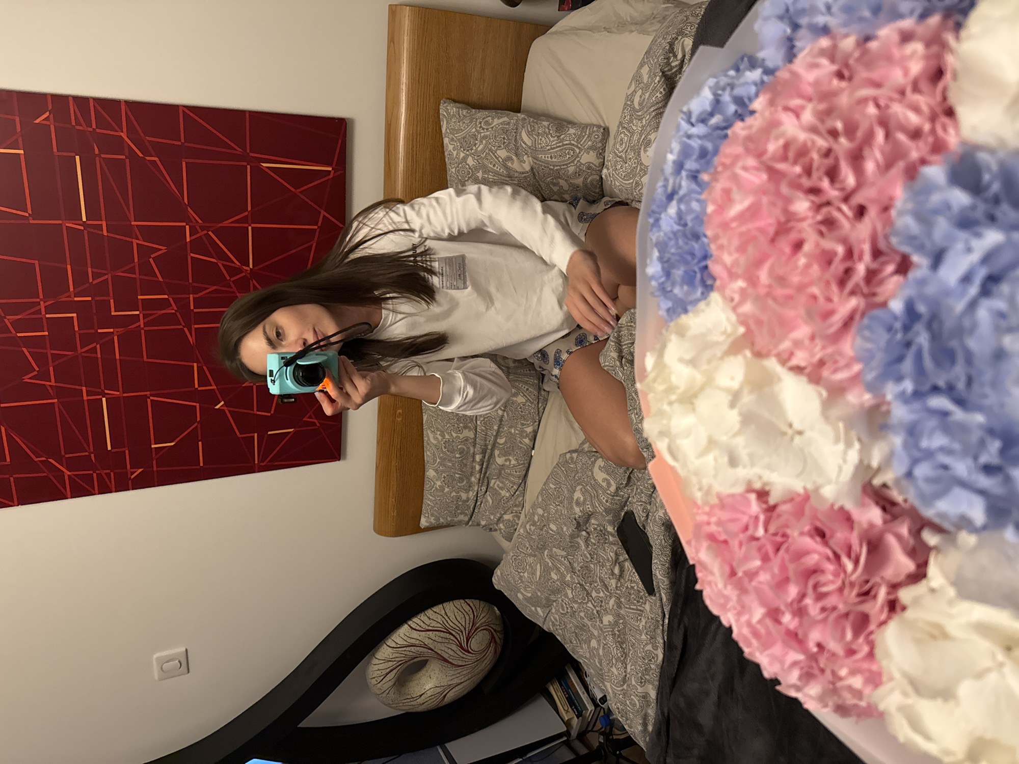 A young woman with long dark hair, wearing a white long-sleeved shirt and patterned shorts, is depicted taking a selfie with a light blue instant camera, likely in a mirror. She is positioned on a bed with a wooden headboard and light-colored bedding adorned with a paisley pattern. Her gaze is directed towards the camera she holds.
On the wall behind her, a large abstract painting features a dark red background with geometric lines in red and orange. Below the painting, a white electrical outlet is visible. Further left, a decorative object resembling a white, donut-shaped sculpture with red swirling patterns rests within a dark, curved base.
To the right side of the image, a large bouquet or arrangement of artificial flowers, predominantly in shades of pink, light blue, and white, is prominently displayed. The scene suggests an indoor setting, likely a bedroom, with artificial lighting. The text on the woman's shirt is not clearly legible.