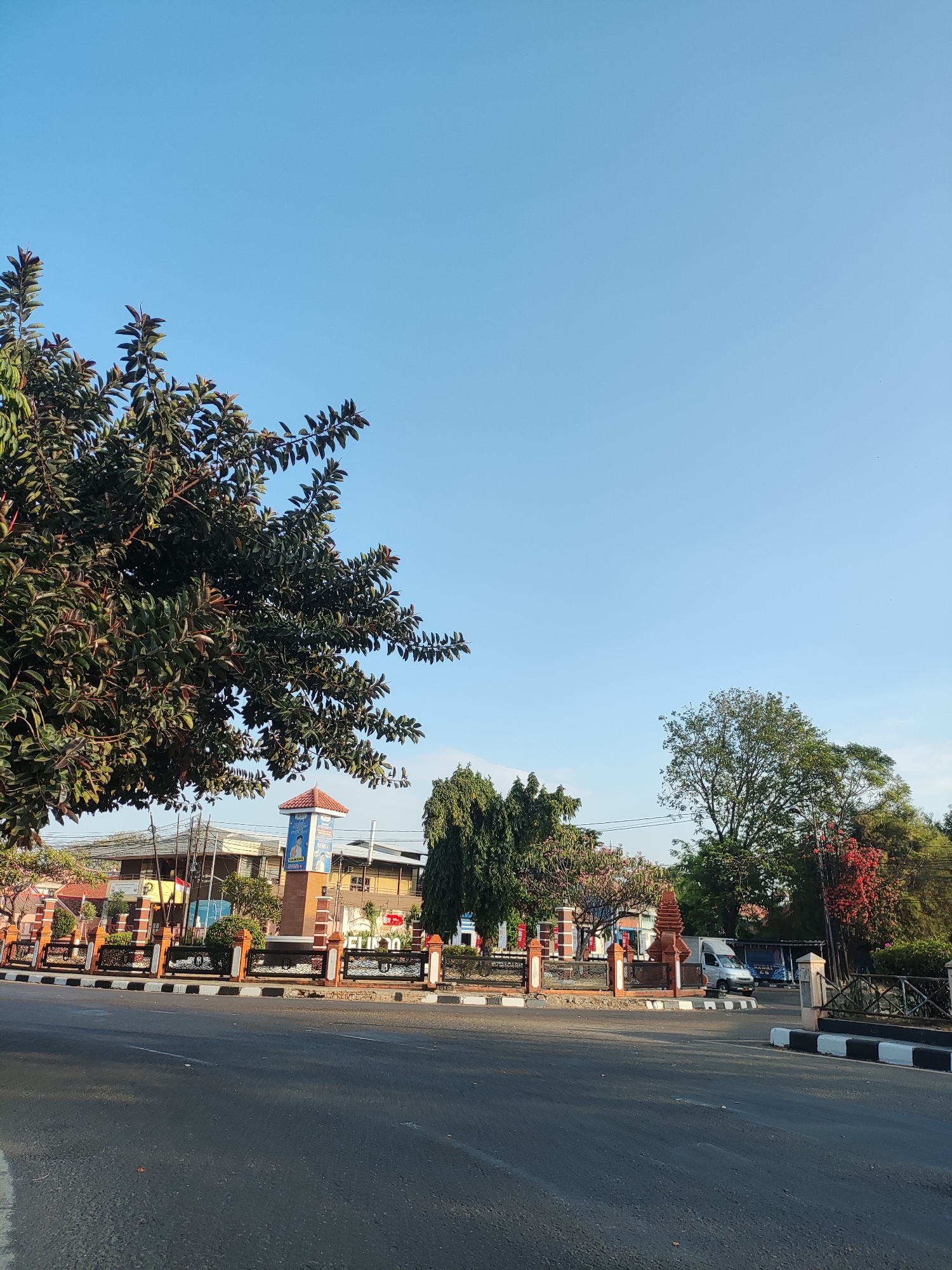 The image shows a street scene in Cirebon, Indonesia. In the foreground, there is an asphalt road with white markings. To the left of the road, there is a large tree with dark green leaves. In the background, there is a monument with an orange roof and a blue banner. The monument is surrounded by a decorative fence with orange pillars and black metal railings. Beyond the monument, there are buildings and more trees. A light blue truck and a darker blue truck are parked in front of a building on the right side of the image. The sky is clear and blue. The scene appears to be set in a public space within the city, possibly a town square or a busy intersection, given the presence of the monument and the road infrastructure.