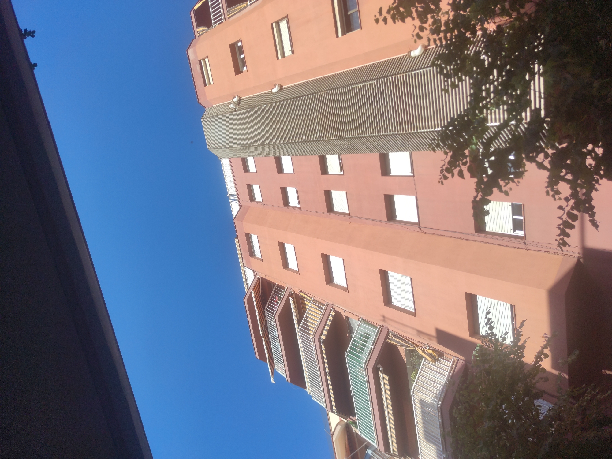 The image depicts a tall, multi-story building in Unknown, Unknown, viewed from a low angle. The building is primarily salmon-colored, with rows of rectangular windows. Some windows have white shades or blinds. A section of the building features vertical, gray louvers. The building also has balconies with varying designs, some enclosed with what appears to be white metal bars and green tinted glass. A deep blue sky serves as the backdrop. In the foreground, the edge of a dark gray structure is visible to the left, and the branches and leaves of a tree partially obscure the building on the right side. The light suggests a sunny day.