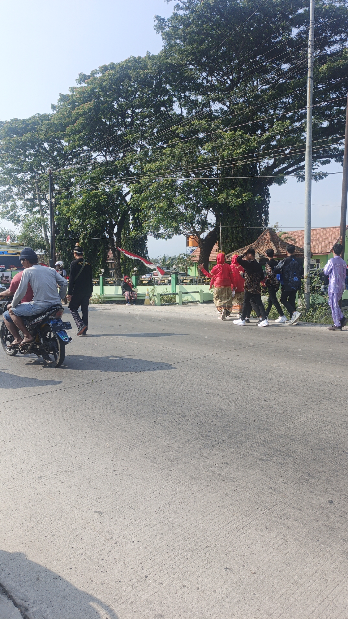In the city of Sumengko, Indonesia, a group of people are seen on a street with a large tree canopy overhead and a clear blue sky. A motorcycle with two occupants is in the foreground, moving from left to right. The driver wears a gray long-sleeved shirt and a blue head covering, while the passenger wears a red shirt and blue shorts. To the right, a group of four young people walk in the same direction, dressed in various casual outfits. Another individual in a red headscarf and traditional skirt stands near the road. Further to the right, a person in a purple garment walks away from the camera. The background shows buildings with tiled roofs and a green fence, suggesting a residential or public area. The road surface is asphalt with visible tire marks.