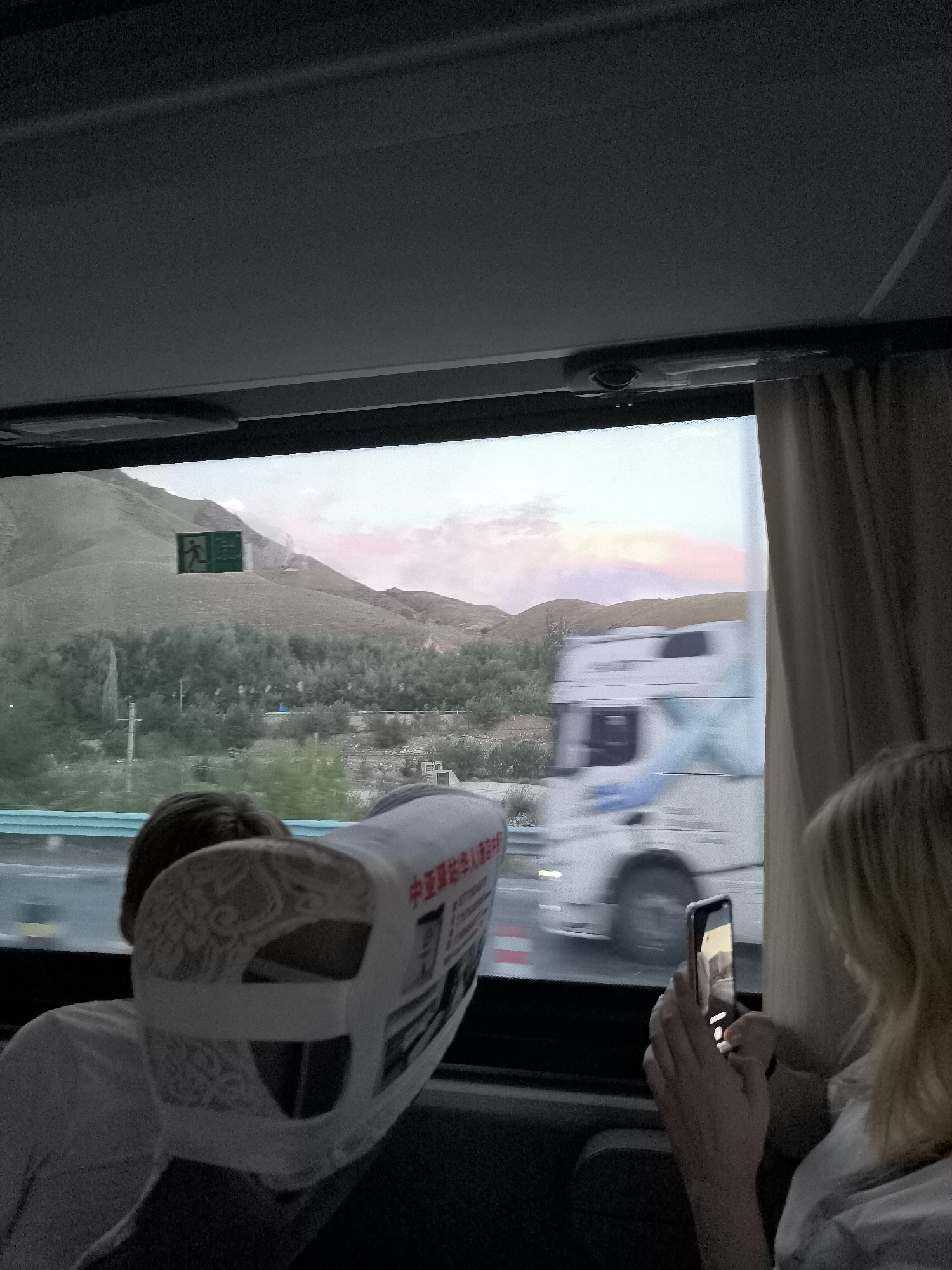 An outdoor scene, as viewed from the interior of a vehicle, captures a truck driving on a highway with a mountainous landscape in the background under a twilight sky. In the foreground, two individuals are partially visible. On the left, the back of a person's head is seen, with the headrest of their seat in front of them. On the right, a person with blonde hair is holding a smartphone, apparently taking a picture or video. The truck is white with blue accents and appears to be in motion, as indicated by motion blur. A green sign with a pedestrian symbol and Chinese characters is visible on the side of the road in the mid-ground. The surrounding environment includes sparse vegetation and a hilly terrain. The lighting suggests it is late evening or early morning. The location is identified as the Guozigou Pasture in China.
