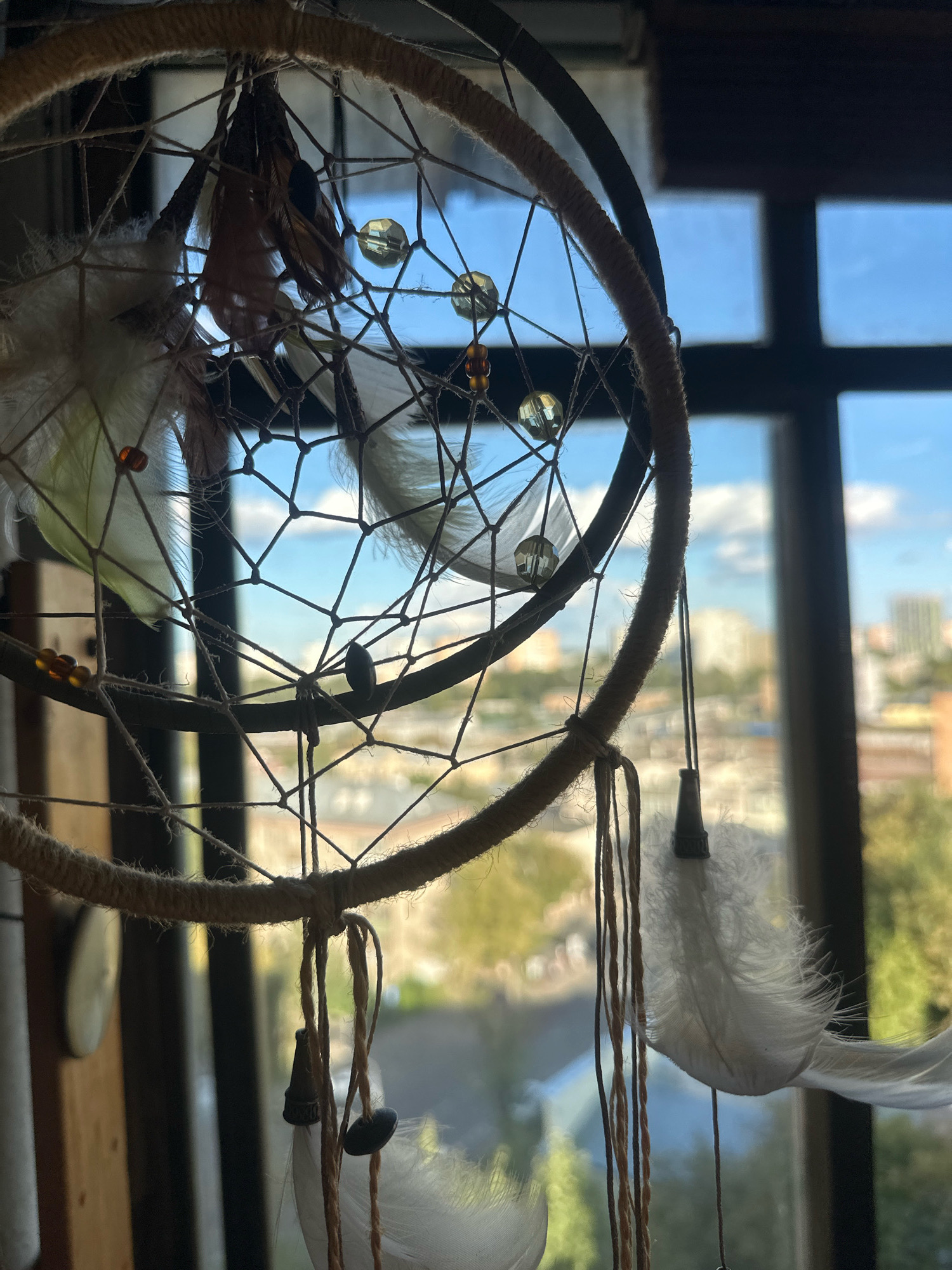 A dreamcatcher hangs in front of a window with a view of a city. The dreamcatcher has a circular frame wrapped in twine, with a web of string stretched across it. Several feathers and beads are attached to the dreamcatcher, including white feathers and amber-colored beads. The window shows a blurred background of buildings, trees, and a blue sky with clouds, suggesting an urban environment. The scene is brightly lit by natural light coming through the window. No people or actions are depicted. The location is not specified in the image or the provided crops.
