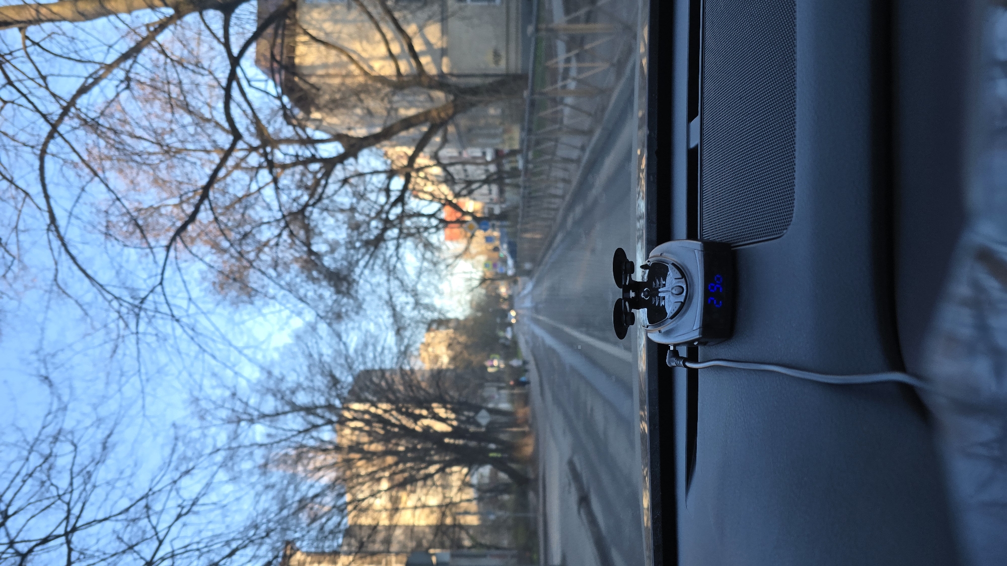 The image is taken from inside a vehicle, looking out the windshield onto a city street. The view includes buildings, bare trees, and a road with lane markings. A small electronic device with a fan and a digital display showing "29.6" is affixed to the interior of the vehicle, likely a temperature or humidity sensor. The location is inferred to be Kaliningrad, Russia, based on visual characteristics.