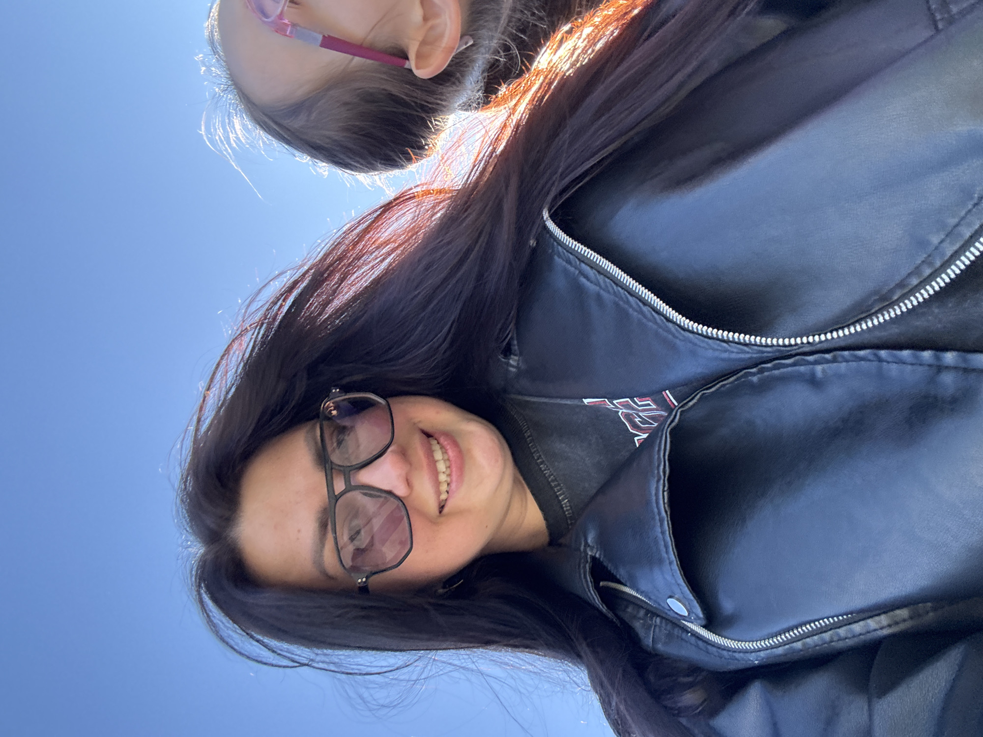 The image captures a low-angle, upward-facing perspective of two individuals against a bright, clear blue sky. In the foreground and appearing on the left side of the frame, an adult woman with long, dark hair is smiling broadly, showing her teeth. She wears dark-rimmed glasses, which reflect some light, and a black jacket, likely leather or faux leather, featuring a prominent zipper and snap fasteners. A dark t-shirt with a partially visible red and white graphic is worn underneath the jacket. The sunlight brightly illuminates her hair, creating warm, reddish highlights. Behind and above her, mostly out of frame, the top of a child's head is visible, sporting pink-framed glasses. The child's face is not discernible. The scene suggests a casual outdoor moment on a sunny day, possibly a selfie, with the adult expressing happiness or contentment. There are no specific location cues other than the open sky. No legible text is visible in the image.