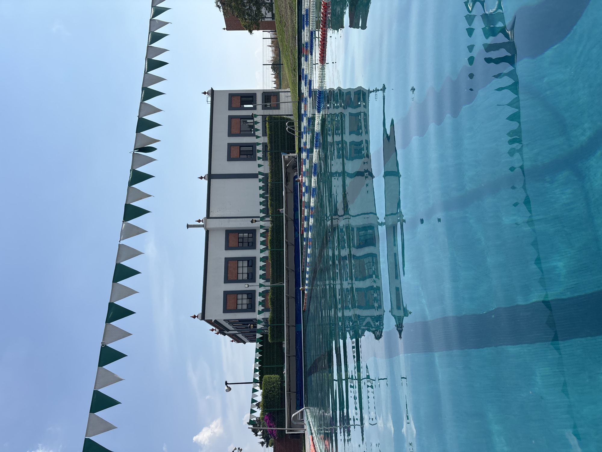 The image shows a building reflected in the water of a swimming pool, with triangular green and white flags strung along the side. The building is white with dark framed windows and is located in San Andrés Cholula, Mexico. Swimming pool lane dividers run alongside the building and into the pool. The water surface is rippled, distorting the reflection. The sky is blue with some wisps of clouds visible to the left. A light pole and vegetation are also visible near the building.