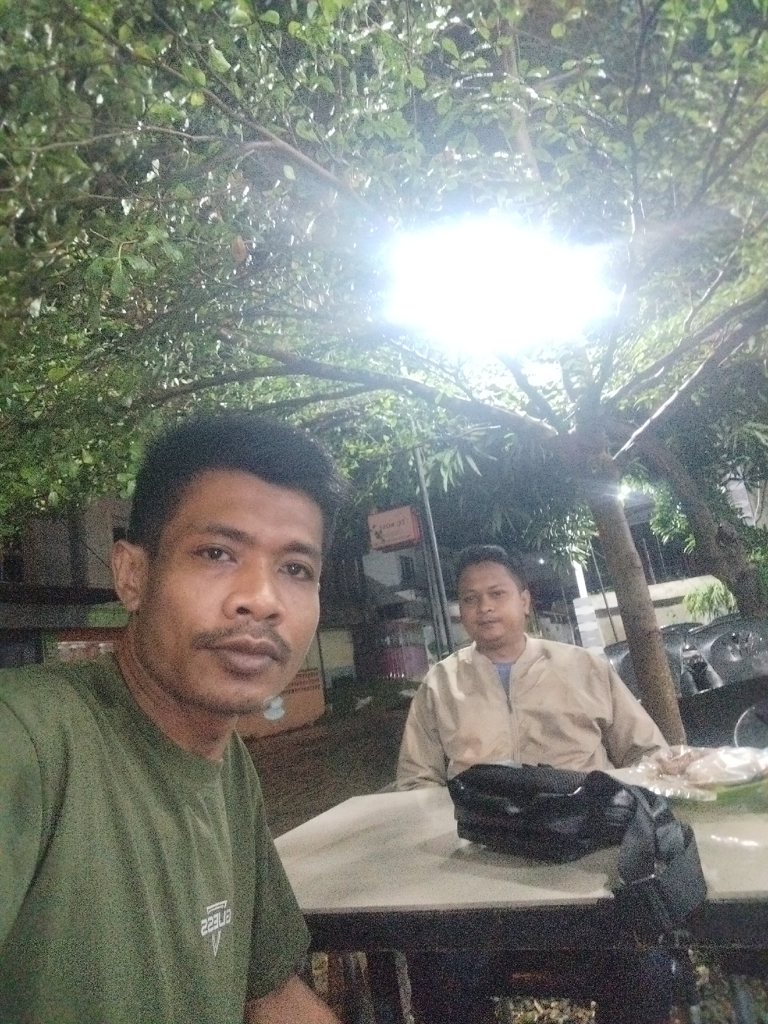 Two men are seated at an outdoor table at night. The man on the left wears a green t-shirt, while the man on the right wears a light tan jacket. A black bag sits on the table between them. The table appears to be made of stone. Above them is a tree with bright lights shining through its branches. In the background are buildings and streetlights. The location is Lhokseumawe, Indonesia.