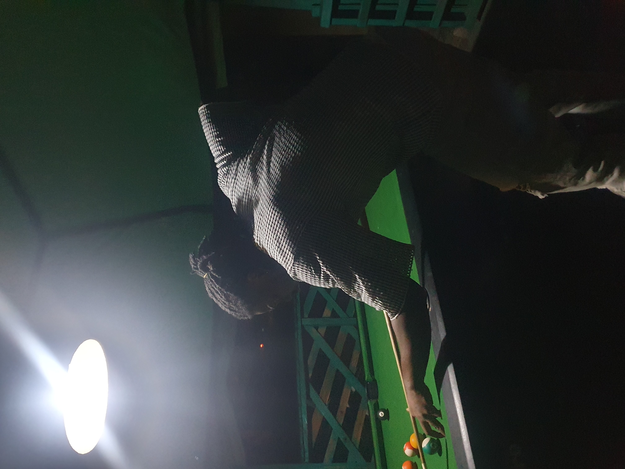 A person with dreadlocks is playing pool in Lokoja, Nigeria. The person is wearing a patterned shirt and light-colored pants. They are leaning over a green pool table, holding a pool cue. Several billiard balls are visible on the table, including an 8-ball. The scene is illuminated by a bright overhead light. In the background, there is a green wooden fence and a dark sky.