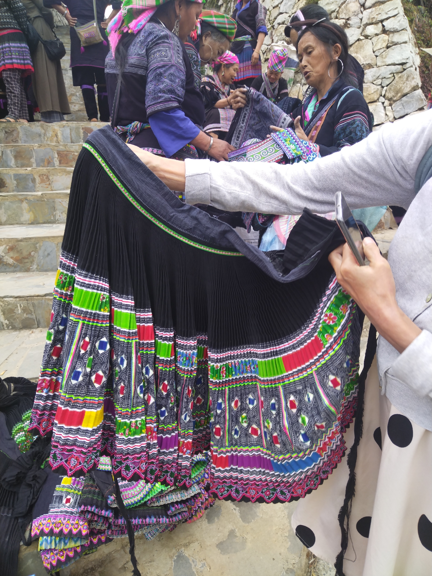 The image captures an outdoor scene featuring several women, primarily dressed in colorful traditional clothing, engaged in a display or sale of textiles. In the foreground, a black, heavily pleated skirt adorned with vibrant patterns in red, green, blue, purple, and white is held up, its intricate geometric and wavy designs visible. The person holding the skirt is wearing modern grey and white attire and is also holding a smartphone.
In the mid-ground and background, multiple women, likely of an indigenous ethnic group, are seen on stone steps and beside a rough-hewn stone wall. They wear elaborate traditional garments, including blouses with patterned sleeves, shoulder bags, and distinctive head wraps in colors like pink, green, and blue. Their clothing features intricate embroidery and patterned fabrics, showcasing rich cultural artistry. One woman with large hoop earrings appears to be conversing or demonstrating another piece of patterned fabric.
The setting suggests a market, village gathering, or a tourist interaction in a mountainous or hillside region, indicated by the stone steps and wall. The activity centers around the viewing or transaction of these traditional textile products. The time of day is clearly daytime, with natural light illuminating the scene. A small, illegible white label or tag is partially visible inside one of the skirts being held. The overall mood is one of focused interaction and cultural display.