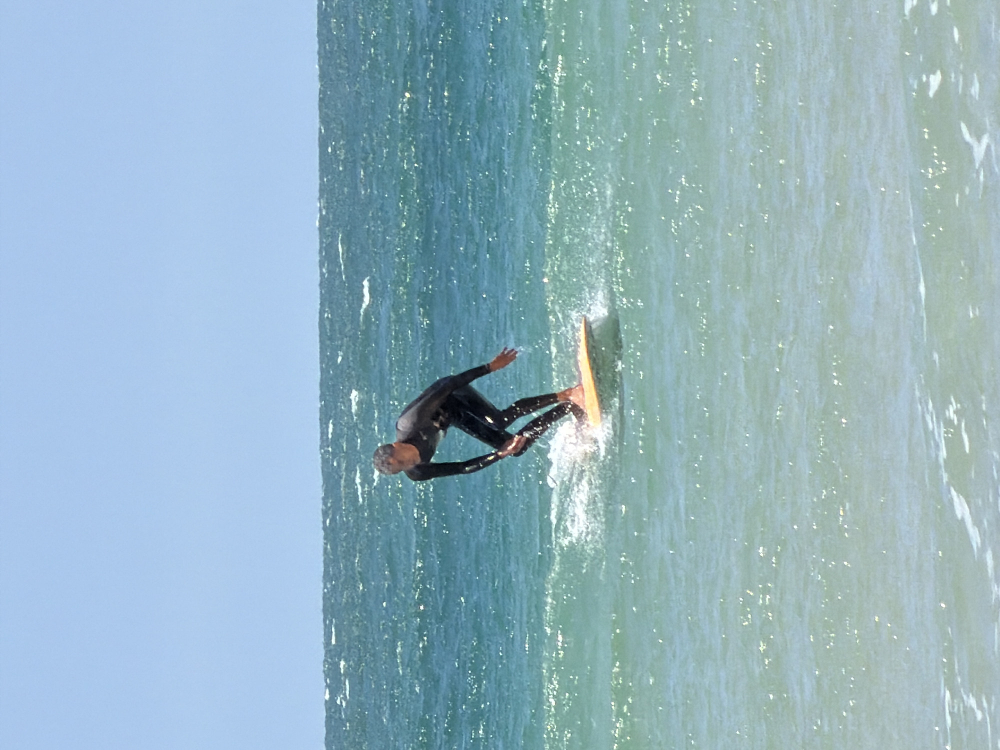 A person wearing a full-body black wetsuit is surfing on a wave. The surfer is positioned on a short, light-colored surfboard, with splashes of water visible around the board. The background consists of the ocean, merging into a light blue sky above. The location is likely a beach in Tel Aviv, Israel.