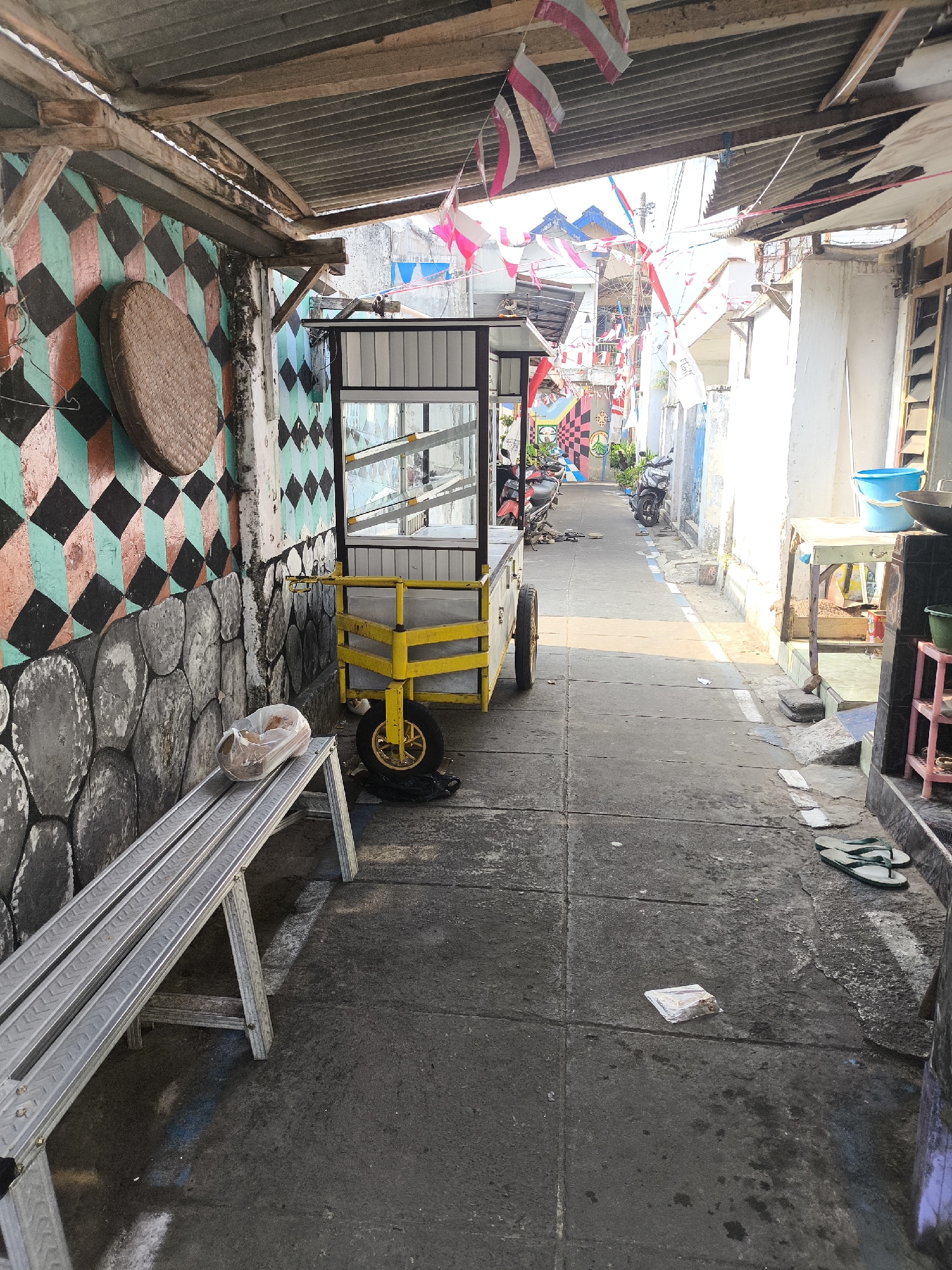 This image depicts a narrow alleyway in Lumajang, Indonesia, likely in a residential or market area, given the presence of a food cart and various domestic items. The alley is paved with concrete and is lined by buildings with corrugated metal roofs. The walls of the buildings are decorated with colorful murals and stone patterns.
In the foreground, a metal bench is placed against a decorated wall. A plastic-wrapped item, possibly food, rests on the bench. A yellow food cart with glass display cases stands to the right of the bench, its single wheel visible. Further down the alley, several motorcycles are parked.
The scene is adorned with numerous Indonesian flags, suggesting a national celebration or event. The overall impression is one of a lively, albeit somewhat weathered, urban environment. The lighting suggests it is daytime, possibly late morning or early afternoon, with sunlight casting shadows on the ground. The presence of flip-flops and a blue bucket implies everyday activity.