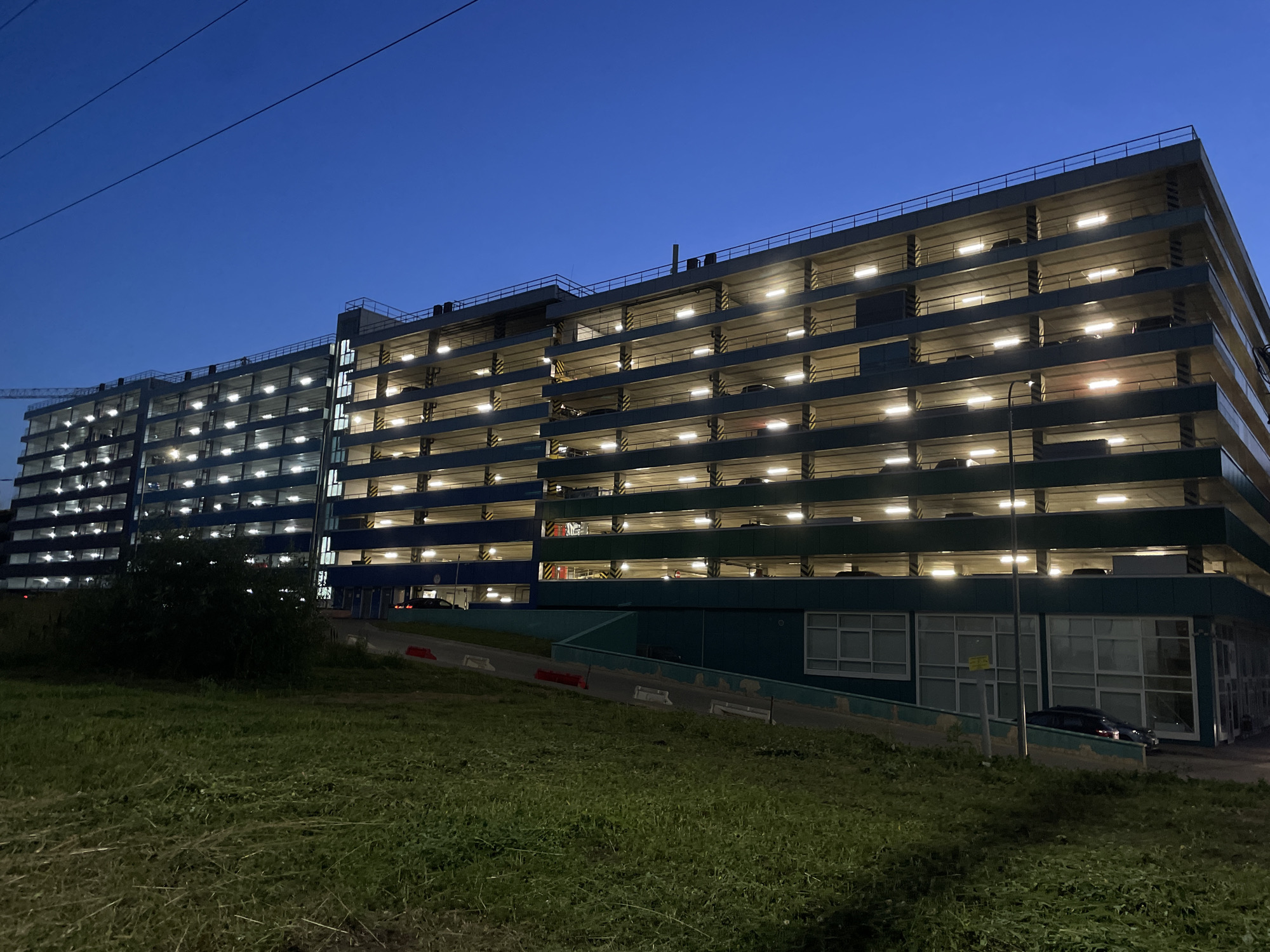 The image depicts an illuminated multi-level parking garage at dusk in Odintsovo, Russia. The building, characterized by its concrete structure and uniformly lit interior levels, is viewed from a slightly elevated grassy embankment. Overhead power lines are visible against a darkening blue sky. The garage has several open levels, each lined with bright lights and filled with parked cars, some of which are discernible as dark silhouettes. The lower section of the building is painted in dark blue and green hues, with large windows on the ground floor. A dark-colored car is parked on the ground level. The foreground consists of a grass-covered slope.