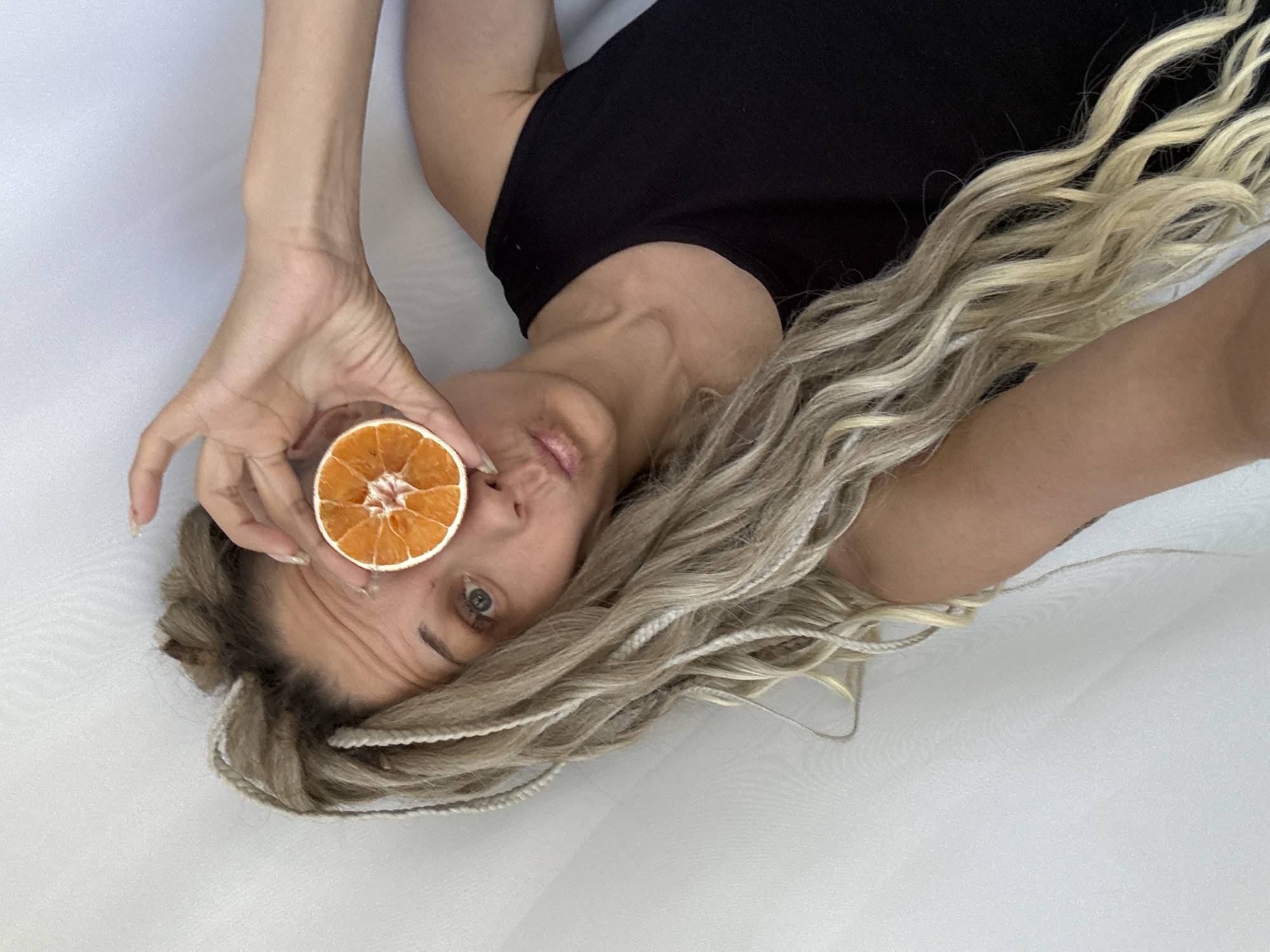 A woman is lying on a white surface, holding a dried orange slice in front of her eye. She is wearing a black top. Her hair is long, blonde, and styled in braids. The background is a plain white surface. There is no visible evidence to determine if this image was taken in Rusanovo, Russia.