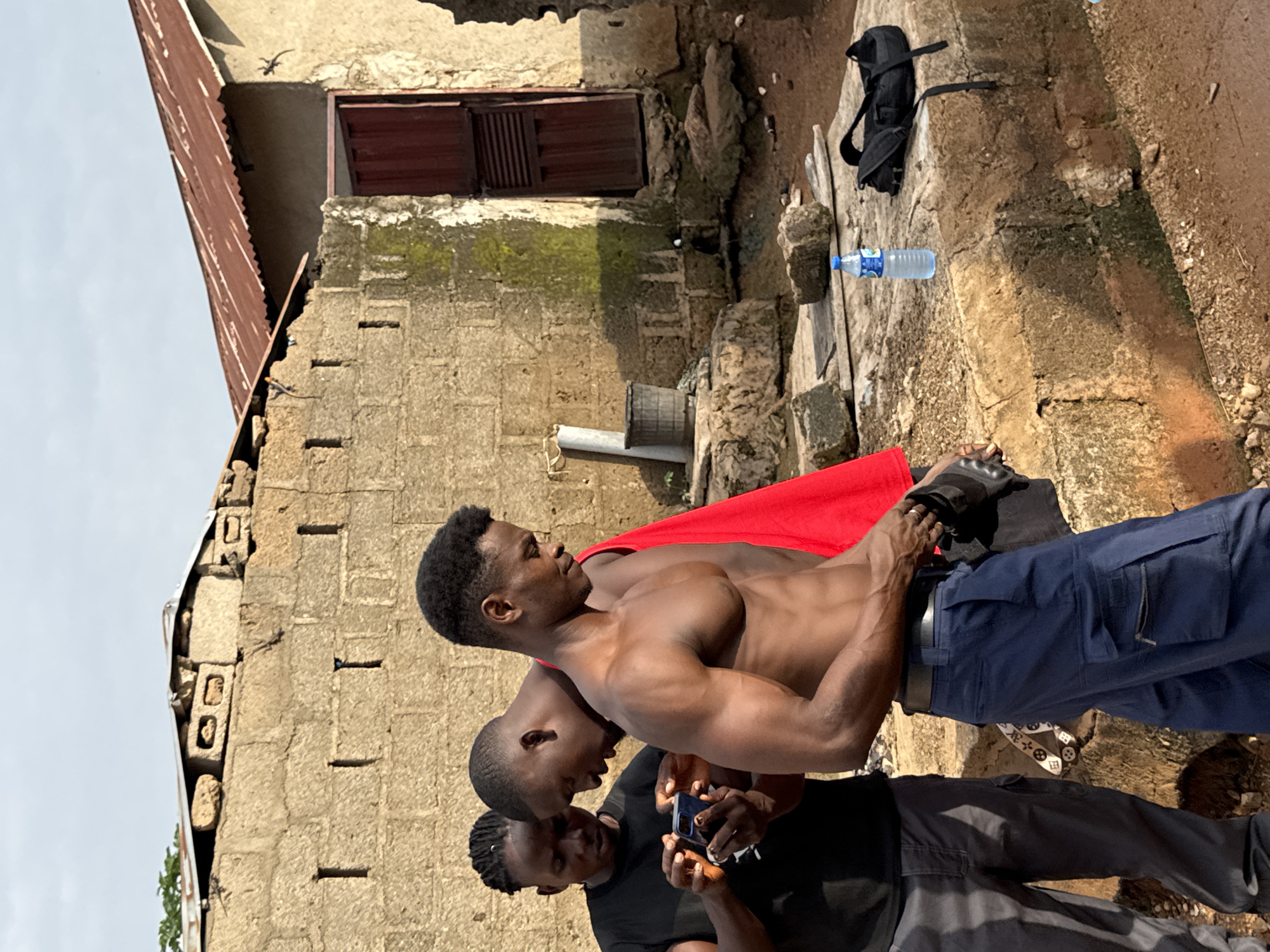 The image shows three young Black men in an outdoor, possibly rural, setting in Akwanga, Nigeria. Two men are in the foreground, and one is slightly behind them. The man in the middle, shirtless and with a muscular physique, is wearing a red cape over his shoulders and blue cargo pants. His gaze is directed to the left. The man on the far left is also shirtless and looking down, holding a smartphone. The third man, mostly obscured, is wearing a black t-shirt and dark pants and appears to be looking at the phone as well.
Behind the men, there is a weathered building constructed with rough-hewn blocks and a corrugated metal roof. A dark wooden door with vertical slats is visible on the building. Patches of green moss grow on the wall. The ground is uneven and appears to be dirt and rocks. A black backpack and a plastic water bottle are placed on a raised stone surface to the right of the men.
The lighting suggests it is daytime, likely late morning or afternoon, with bright sunlight casting shadows. The overall atmosphere appears casual and possibly recreational. There is no visible text in the image.