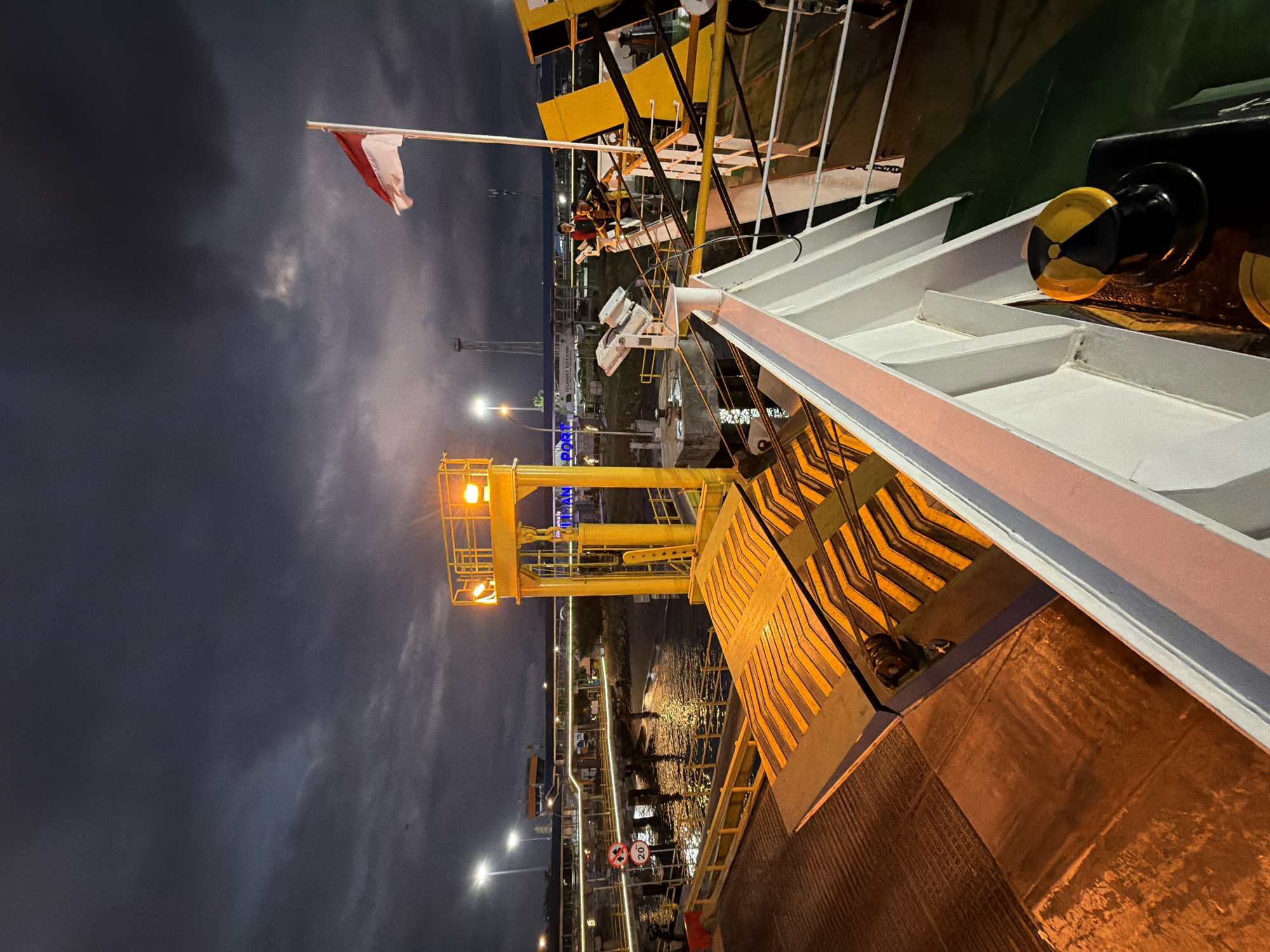 The image depicts a nighttime scene at a port in Melaya, Indonesia, likely a ferry terminal. The sky is overcast and dark, with some streaks of lighter clouds. A red and white Indonesian flag is prominently displayed on a flagpole to the right of the frame, rippling in the wind.
In the foreground, part of a white ship's railing and a black and yellow bollard are visible. Extending from the ship is a large yellow ramp, which appears to be a vehicle ramp for a ferry. This ramp has diagonal black and orange reflective markings. Beyond the ramp, there is a yellow gantry structure with lights.
In the middle ground, the water of the port is visible, reflecting the lights from the shore. A walkway or pier extends into the water, lined with lights. On this pier, a sign with a red circle and a diagonal line with the number "20" inside is visible, indicating a speed limit. Further back, there are buildings and structures associated with the port, including what appears to be a terminal building with the word "PORT" illuminated in blue. Two individuals are standing on a raised platform in the middle distance, possibly port workers.
The overall impression is a functional, active port at night, with a sense of transition and movement. The lighting from the port structures and the ship create dramatic highlights and shadows.