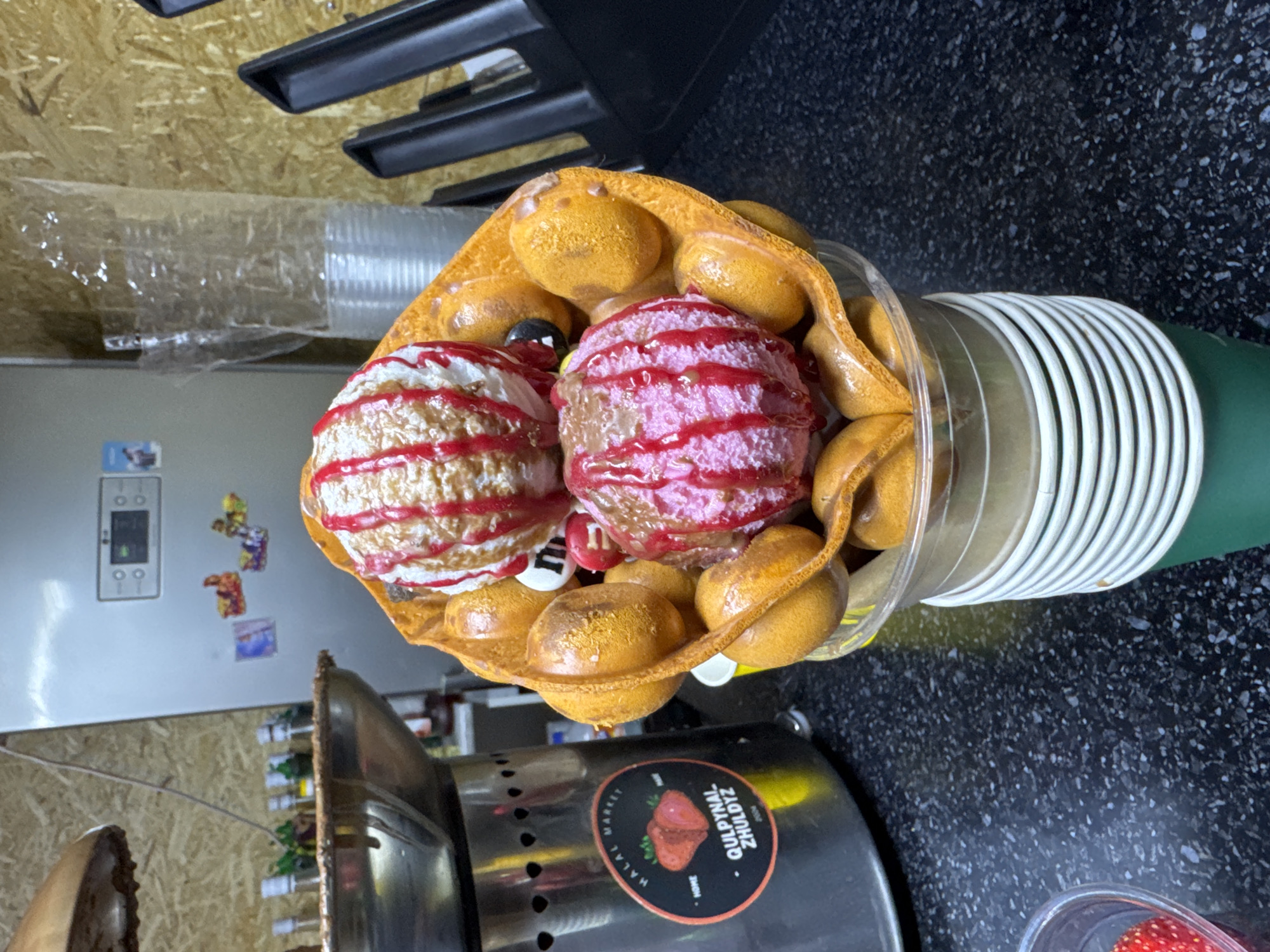The image depicts a close-up, eye-level shot of a bubble waffle ice cream cone dessert. The dessert is presented in a clear plastic cup, stacked on top of a green paper cup. The bubble waffle, a golden-brown color, is filled with two scoops of ice cream. The top scoop is vanilla with red syrup drizzled over it. Below it, a pink scoop of ice cream is also adorned with red syrup. The background shows a wooden-paneled wall with a silver refrigerator on the left, adorned with colorful stickers. To the right of the waffle cone, a stack of clear plastic cups is visible. In the lower left corner, a small plastic container holds red berries, possibly strawberries. The overall scene is likely within a food establishment in Zharbulak, Kazakhstan, given the presence of a "Halal Market" sticker with "Qulpynal Zhuldyz" branding on a stainless steel machine to the left of the dessert.
