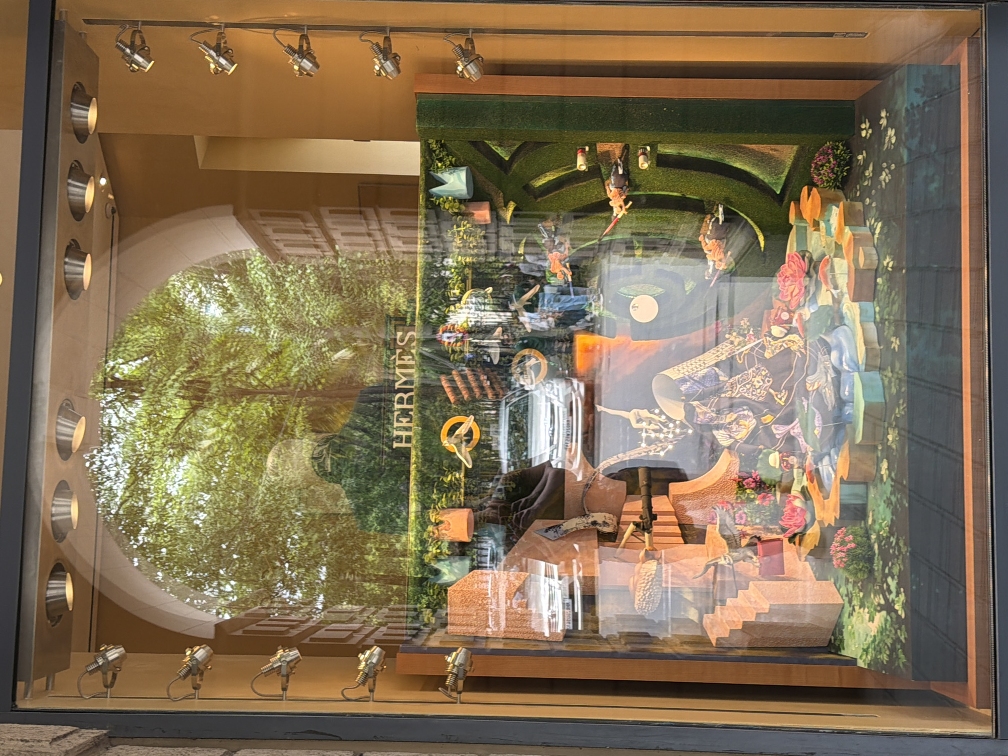 This is a photograph of a luxury storefront window display, likely in Mumbai, India, given the presence of several white sedans, a common car model in India. The window is framed by dark metal and wood accents. A series of industrial-style spotlights are mounted on the wall above and to the sides of the window. The display itself is elaborate and features a diorama-like scene.
The background of the window reflects trees and buildings, suggesting it faces a street. The main display area is divided into sections. The top section displays the "HERMÈS" logo prominently. Below this, there is a scene with what appears to be a stylized, abstract landscape. Small, decorative elements are scattered throughout.
In the foreground of the display, there are several figures that appear to be dressed in elaborate, traditional-looking attire, possibly inspired by Indian or South Asian culture. They are posed in a theatrical manner amongst various colorful and textured elements, including what looks like a lotus flower and water features. The overall impression is one of a fantasy or artistic representation.
There are reflections visible on the glass, including a person holding a camera, indicating the photograph was taken from outside the store. The lighting within the display is strategic, highlighting the products and the artistic elements. The colors are rich and varied, with a focus on greens, oranges, blues, and pinks. The overall scene is detailed and designed to attract attention.