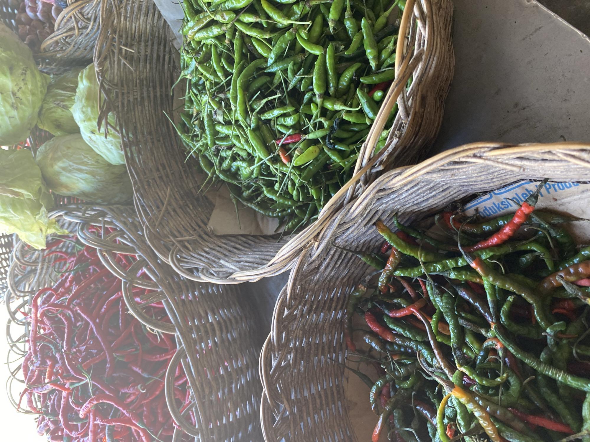 The image shows an overhead shot of four woven baskets filled with chilies and some cabbage. In the top left, a basket is overflowing with long, red chilies. To its right, another basket contains several heads of light green cabbage. Below these, a basket on the left is filled with a mix of green and red chilies, some appearing slightly dried or withered. The bottom right basket is packed with numerous small, green chilies. The baskets are placed on a flat surface, possibly a market stall, and the overall lighting suggests an outdoor or well-lit indoor setting.