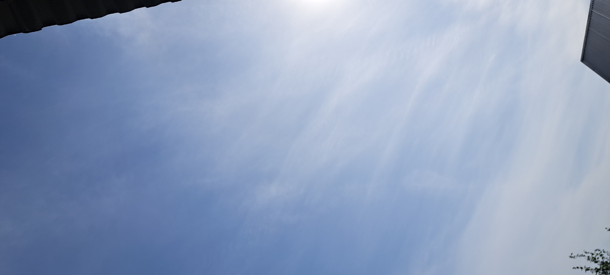 The image shows a bright blue sky with thin, wispy clouds. The sun is partially visible on the right side of the frame, casting a glare. The top right corner of the image is occupied by the dark, angled edge of a roof, likely part of a building. In the bottom left corner, the top of some foliage, possibly a tree, is visible. In the bottom right corner, the peak of a dark, sloped roof is present, likely another building. The scene appears to be outdoors, possibly in a suburban or rural area, given the presence of roofs and foliage. The location of Азьмушкино, Russia, is not visually represented in the image itself, as there are no specific landmarks or identifying features of that particular location.