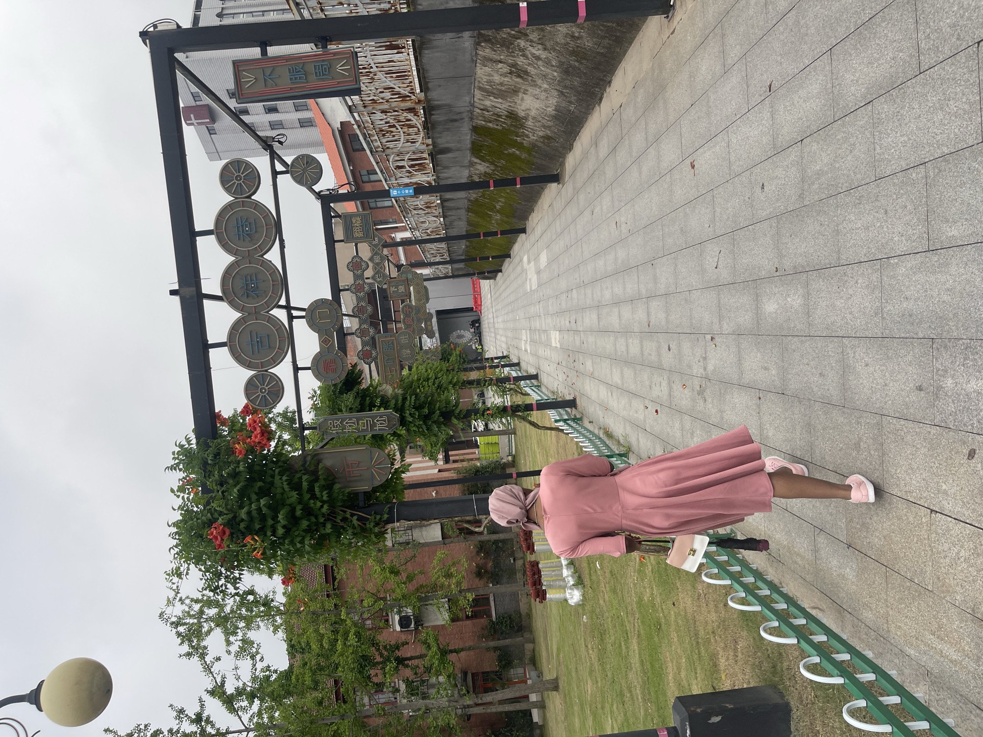 The image depicts an outdoor pedestrian walkway in the city of Unknown, Unknown, under an overcast, light grey sky. A person, viewed from behind, walks away from the camera on a path paved with rectangular light grey tiles. The individual has dark skin and is wearing a solid pink long-sleeved dress with a flared skirt, a matching pink head covering, and light pink sneakers with white soles. They carry a two-toned handbag, light-colored on top and dark pink/brown on the bottom, along with a folded dark umbrella in their right hand.
To the immediate left of the path is a manicured green grass lawn, bordered by a low green fence with white arched top sections. Beyond the lawn, several deciduous trees with green foliage stand in front of a multi-story red brick building featuring multiple windows and visible air conditioning units. Several red fire suppression pipes are positioned in front of the brick building. A spherical yellow lamp fixture is visible in the upper left corner.
To the right of the path, a concrete retaining wall, partially covered in green moss, supports a rusty, ornate metal railing. Further down the path, a light-colored multi-story building with a red cross symbol on its upper facade is visible. Another building with a red-tiled roof is also seen in the background.
An overhead dark metal framework spans the walkway, creating an archway or canopy. This structure is adorned with numerous circular and rectangular decorative panels, many displaying Chinese characters and symbols. Climbing vines bearing clusters of bright orange trumpet-shaped flowers are intertwined around the posts and crossbeams of the framework. The lower sections of the vertical support posts are wrapped with pink tape or ribbon.