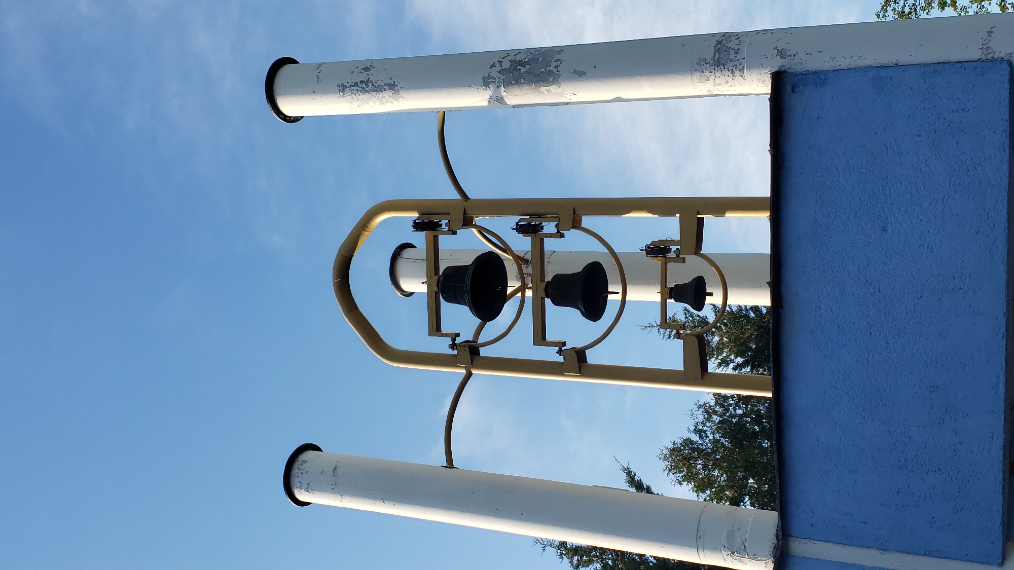 The image shows a carillon, a musical instrument composed of a series of bells, housed within a modern structure. The carillon features at least four dark-colored bells arranged vertically within a yellow metal frame. Two large white cylindrical structures, possibly part of the carillon's support or facade, flank the central frame. The structure is positioned above a blue, textured surface, likely a building facade. Green trees are visible in the background, and the sky above is a clear blue with sparse clouds. This carillon is located at the main entrance of the Slovak Technical University (STU) - Faculty of Civil Engineering in Prešov, Slovakia.