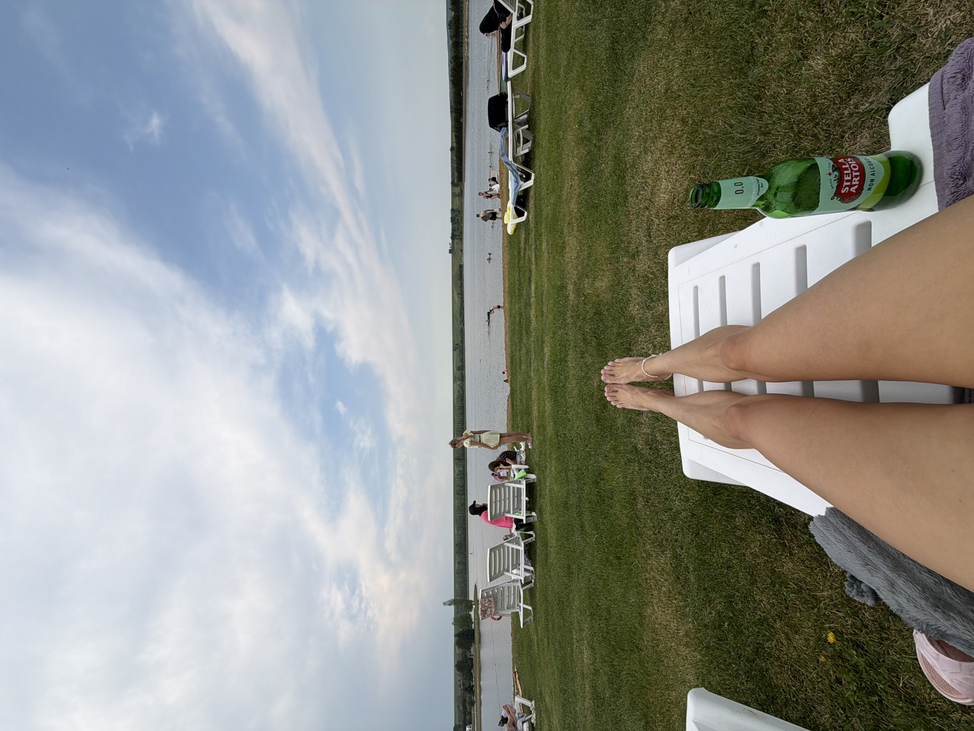 In Yemelyanovo, Russia, a person's legs are stretched out on a white lounge chair, facing a lake with people swimming and sunbathing in the background. A green bottle of Stella Artois non-alcoholic beer rests on the chair next to the person's legs. The scene is set on a grassy area adjacent to the lake, under a partly cloudy sky.