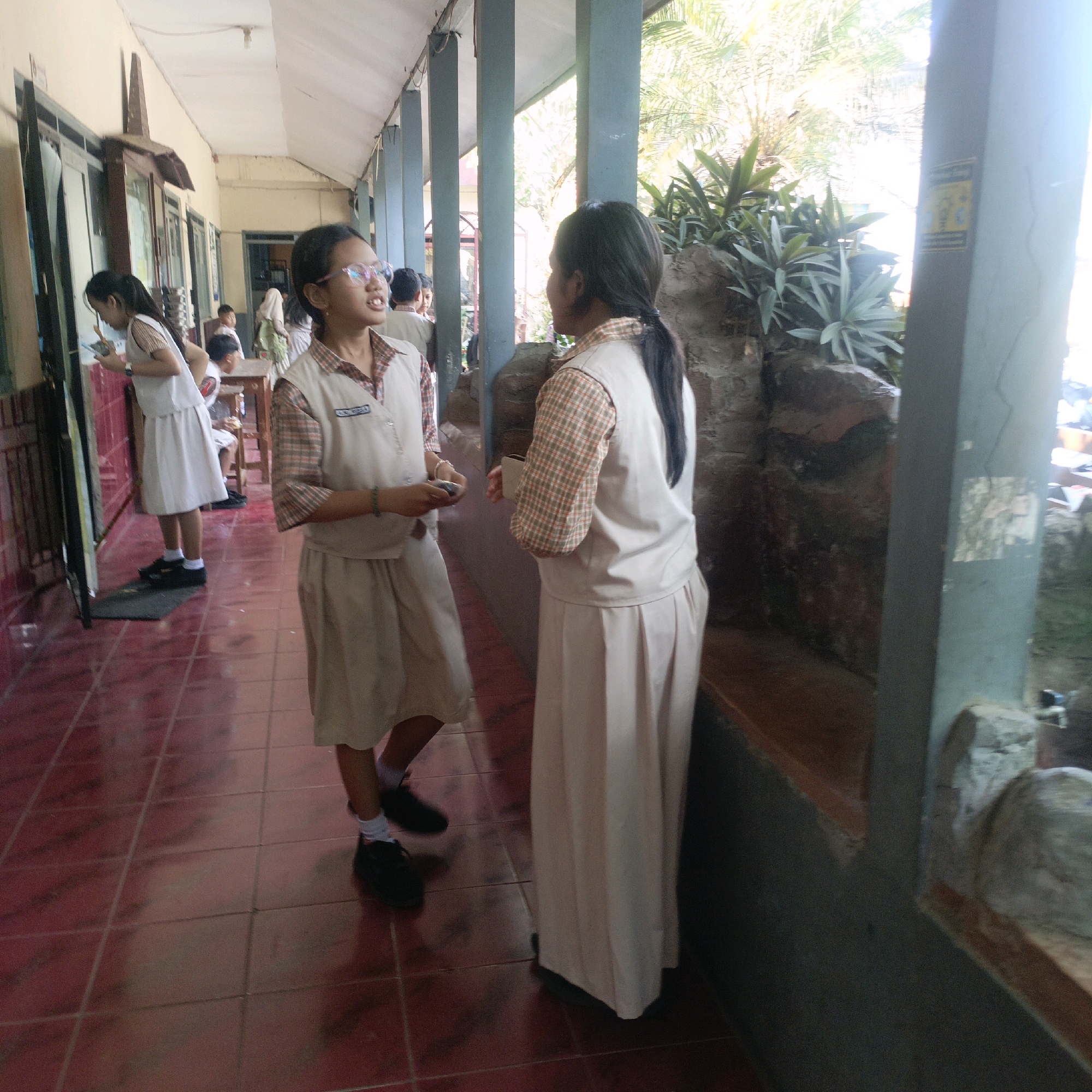 The image shows an outdoor walkway with a tiled red floor and a covered walkway overhead, likely at a school in Salatiga, Indonesia. Two young girls in school uniforms are interacting. The girl on the left, wearing glasses and a white skirt and vest over a plaid shirt, holds what appears to be a phone. The girl on the right, also in a similar uniform with a skirt and vest over a plaid shirt, is facing away from the camera and holding a small object. In the background, other students and a teacher are visible, and there is greenery and rockwork along the side of the walkway. The overall lighting suggests it is daytime.