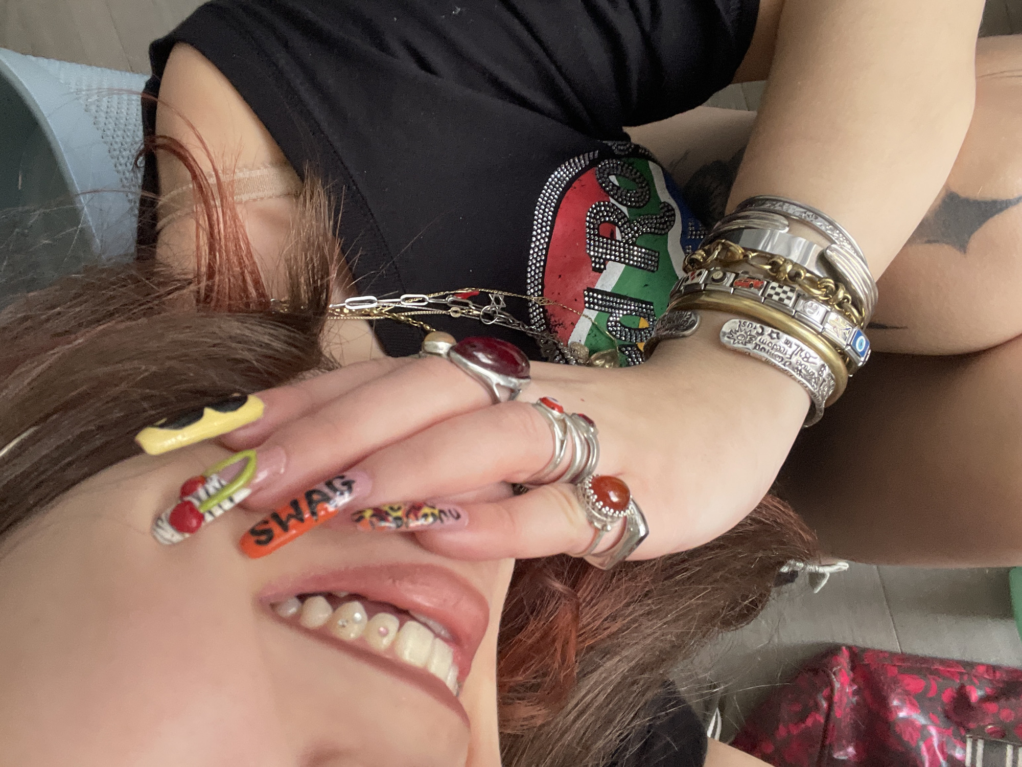 A close-up, high-angle shot captures a person lying down indoors, displaying a distinctive personal style. The subject, likely female, has reddish-brown hair and is smiling broadly with their mouth open, revealing teeth adorned with small, light-colored embellishments, possibly pearls or tiny gems. One hand is raised, partially covering the upper face and showcasing an array of elaborate accessories.
The nails are long and feature intricate designs, including yellow sunglasses on one nail, a green and white sneaker on another, and the word "SWAG" in orange letters on a third. Other nails show animal print or flame-like patterns. Numerous rings adorn the fingers, some featuring prominent red or orange cabochon stones set in silver bands.
The subject wears multiple layered necklaces, including delicate gold chains and thicker silver chains, some with small charms. The wrist is heavily stacked with bracelets of various styles: wide silver cuffs (one resembling a fork handle), gold chains, a charm bracelet with small squares (including a red car and a checkered flag), and an engraved silver bangle that partially reads "...Freedom Trust..." and features a small blue evil eye bead.
The person is wearing a black t-shirt with a vibrant, embellished graphic design visible on the chest, incorporating colors like red, green, and blue, and detailed with sparkling rhinestones. Part of a tattoo, possibly on the arm or upper leg, is visible in the background, appearing dark against the skin. The setting appears to be indoors with a light-colored floor and a blue mat or cushion beneath the head. A red bag with a dark floral pattern is partially visible in the bottom right corner. The overall mood is relaxed and expressive.