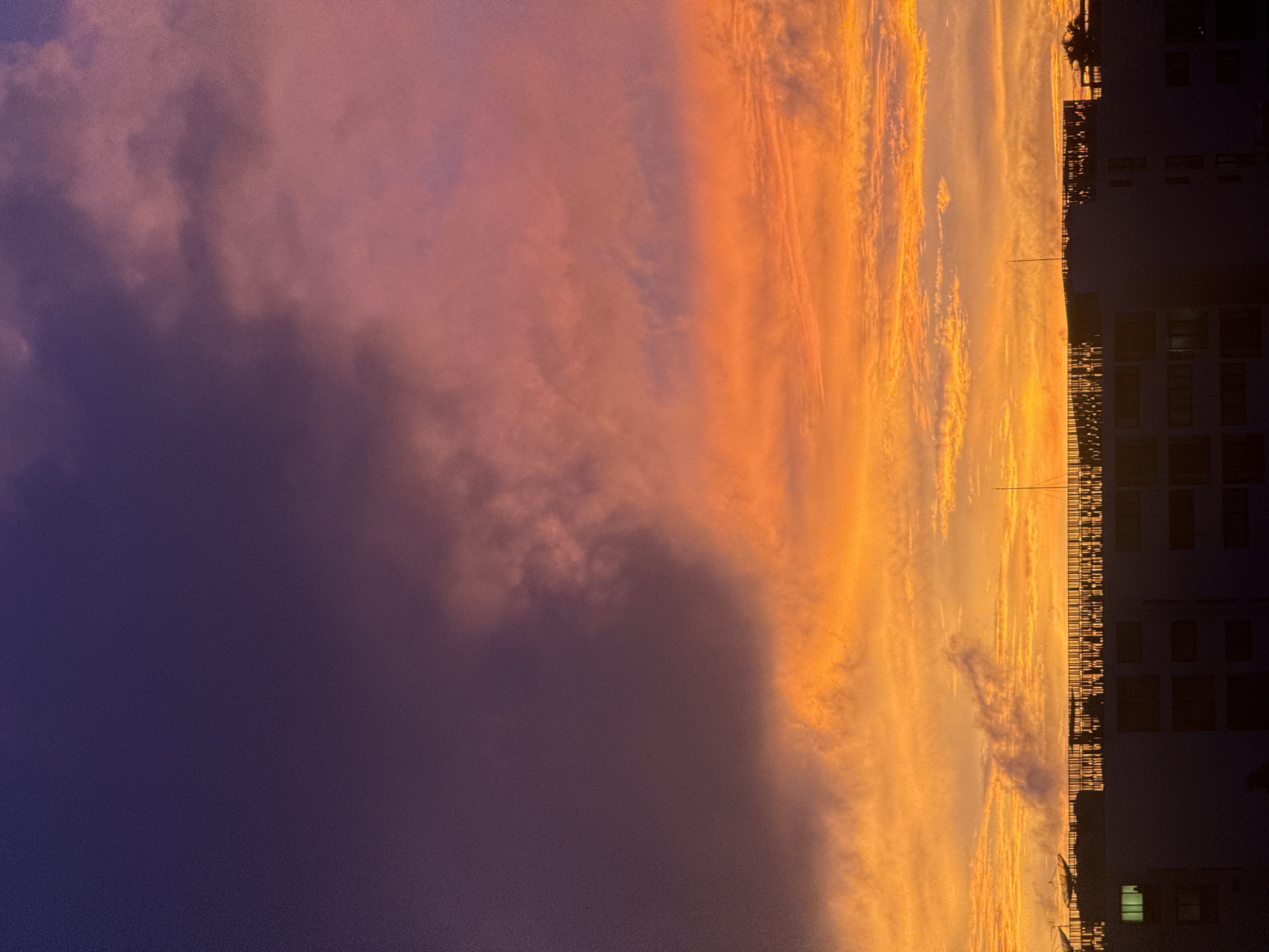 The image depicts a dramatic sunset over Karon, Thailand. The sky is ablaze with vibrant hues of orange, pink, and purple, with heavy, textured clouds dominating the upper portion. Below the colorful sky, the silhouette of buildings and rooftops is visible, providing a stark contrast to the celestial display. The architecture appears modern, with several satellite dishes mounted on one of the structures. The scene is set at dusk, with the city lights just beginning to emerge on the buildings. The overall impression is one of natural beauty and urban life intertwined.