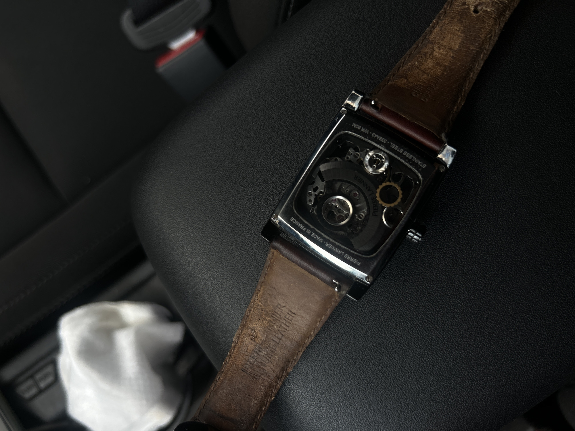 The media file displays a close-up overhead view of the back of a rectangular watch with a brown leather strap, resting on a dark, textured surface inside what appears to be a car interior. The watch is the primary subject and is in sharp focus, while the background is softly blurred.
The watch features a silver-toned, rectangular case with a transparent back, revealing the intricate mechanical gears and components of its movement. Visible text engraved on the case back includes "STAINLESS STEEL - 338A43 - WR 50M" and "PIERRE LANNIER - MADE IN FRANCE," with "PIERRE LANNIER" also visible on the movement itself. The worn brown leather strap is attached to the watch case, showing creases and discoloration from use. Text stamped on the underside of the strap reads "GENUINE LEATHER."
The dark background exhibits a textured surface, likely leather or a synthetic material, characteristic of a car seat or console. In the upper right background, a blurred black seatbelt buckle with a red release button is partially visible. Towards the bottom left, a crumpled white object, possibly a tissue or cloth, is also out of focus. The lighting is ambient, reflecting subtly off the watch's metallic case. There are no people visible, and no specific activity is taking place beyond the watch resting in the car.