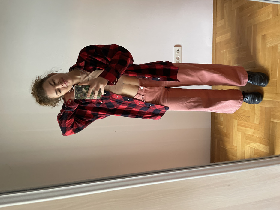 A full-length mirror selfie depicts a female subject standing indoors, in what is identified as Boreti, Montenegro. The subject, possessing curly light brown hair loosely gathered, is smiling gently and holding a smartphone with a green-patterned case in her left hand, directing it towards the mirror to capture her reflection. Her right arm is raised, with the hand positioned behind her head. She is dressed in an open red and black plaid long-sleeved shirt over a beige cropped top that reveals a portion of her midriff, paired with pink wide-leg trousers and black lace-up shoes. A silver chain necklace and rings are visible accessories. The immediate environment includes a plain, light-colored wall, a light-framed mirror occupying the left side of the frame, and light brown herringbone parquet flooring. A white double electrical outlet is mounted on the lower right wall.
