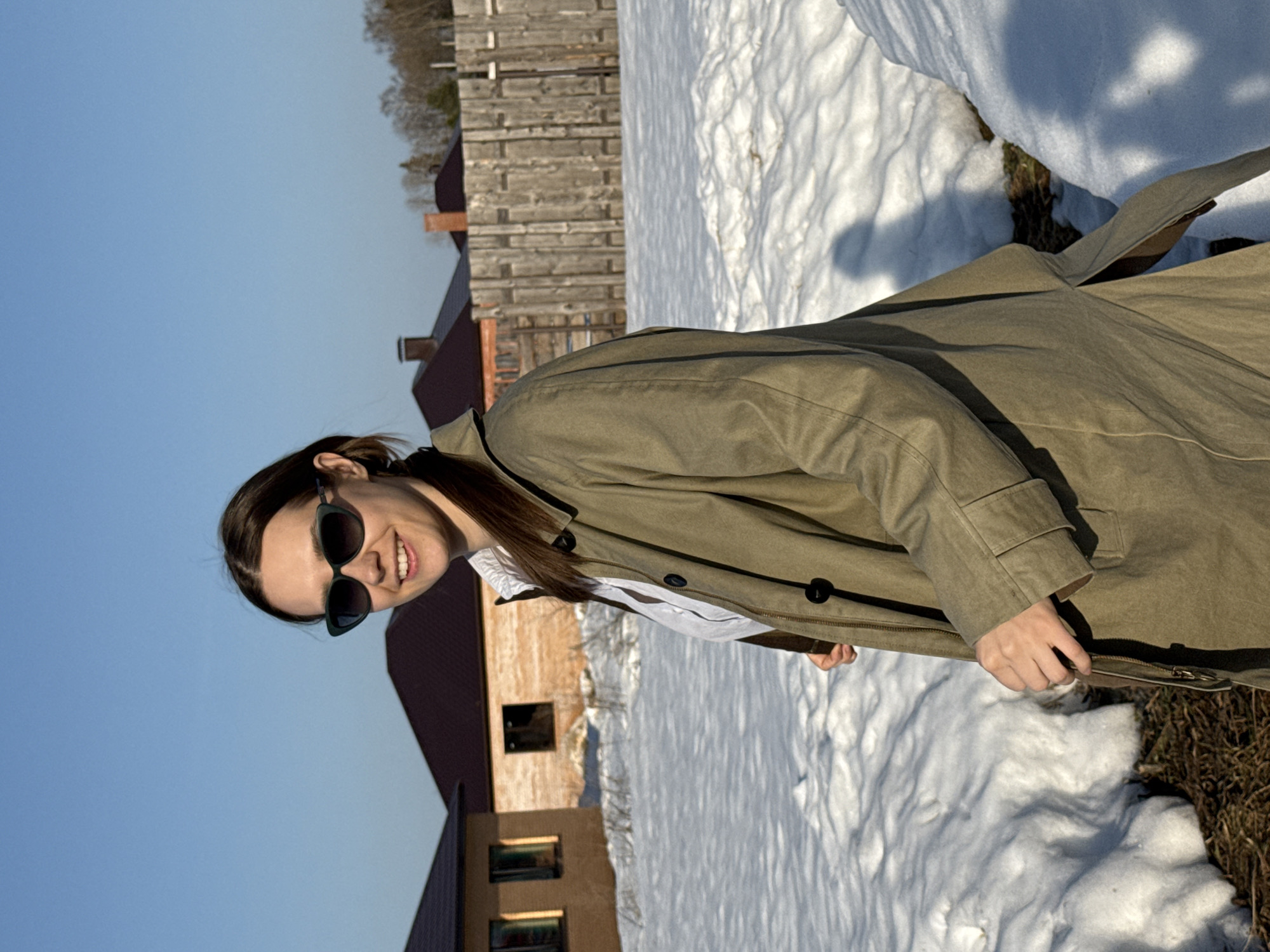 The image displays a female individual outdoors in a snow-covered, residential area of Chaikovsky, Russia. The woman is positioned slightly off-center to the viewer's right, looking directly at the camera with a broad smile. She has dark brown hair, worn back, and is wearing dark green, possibly cat-eye style, sunglasses. Her attire includes an open, olive-green or khaki-colored long coat over a white collared shirt. Her right hand (viewer's left) is positioned near the front of her coat, with a brown strap visible.
The ground is unevenly covered in melting snow, revealing patches of dark earth and dry vegetation. In the background to the viewer's left, two houses with dark brown tiled roofs are partially visible, one featuring two rectangular window openings. To the right, a tall, weathered wooden plank fence stands, separating the immediate area from a line of leafless deciduous trees in the distance. The sky is clear and bright blue, indicating sunny conditions, with discernible shadows cast on the snow from the left, suggesting a low to mid-day sun angle.