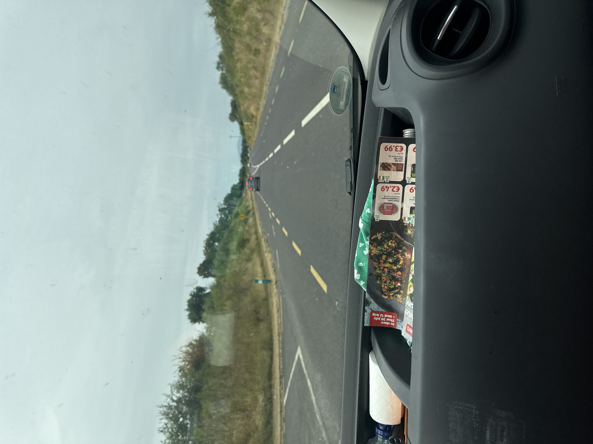 This is a first-person perspective photo from inside a vehicle, likely a van, looking forward through the windshield. The vehicle is traveling on a paved road with a white dashed line separating lanes and solid yellow lines marking the edge. The road is flanked by dry, grassy verges and scattered green trees. The sky is overcast and grey. In the foreground, on the dashboard, a compartment holds a roll of paper towels, a blue bottle, and several flyers or leaflets, one of which displays food items with prices in Euros, specifically "€2.49" and "€3.99". In the middle distance, other vehicles are visible on the road, and a green and white post stands at the side of the road. The overall scene suggests travel through a rural or semi-rural area in Tennypark, Ireland, under cloudy weather conditions. There are no people visible within the frame.